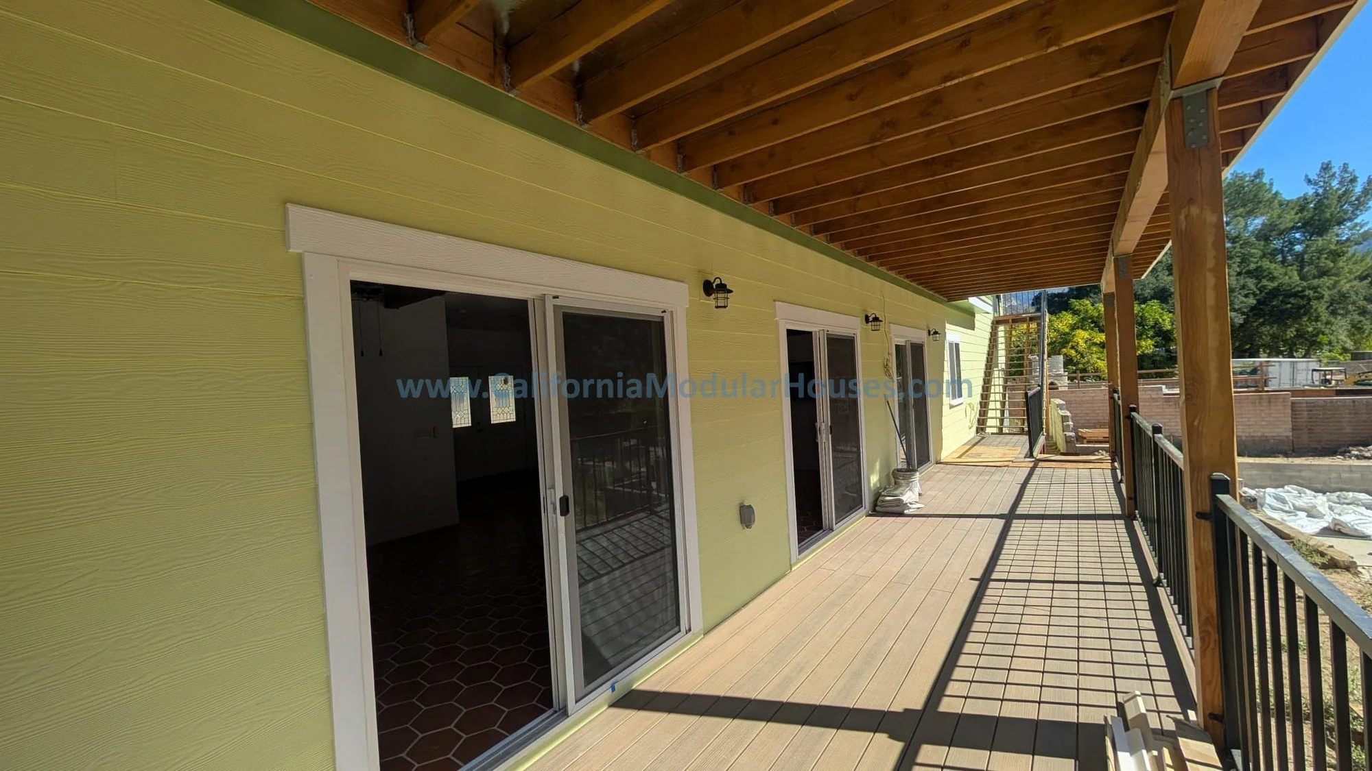 Newly constructed balcony with sliding glass doors, black metal railing, and wooden decking, overlooking a backyard with trees and construction materials.
