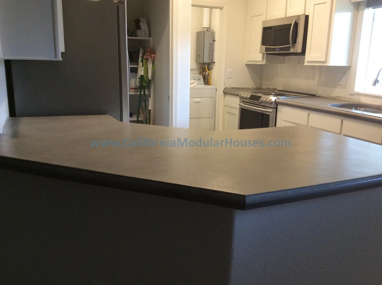 Interior of a kitchen with a beige countertop, white cabinets, a microwave, a stove, a washing machine, and a stainless steel sink near a window. There is a refrigerator and cleaning tools in the background. The website www.CaliforniaModularHouses.co