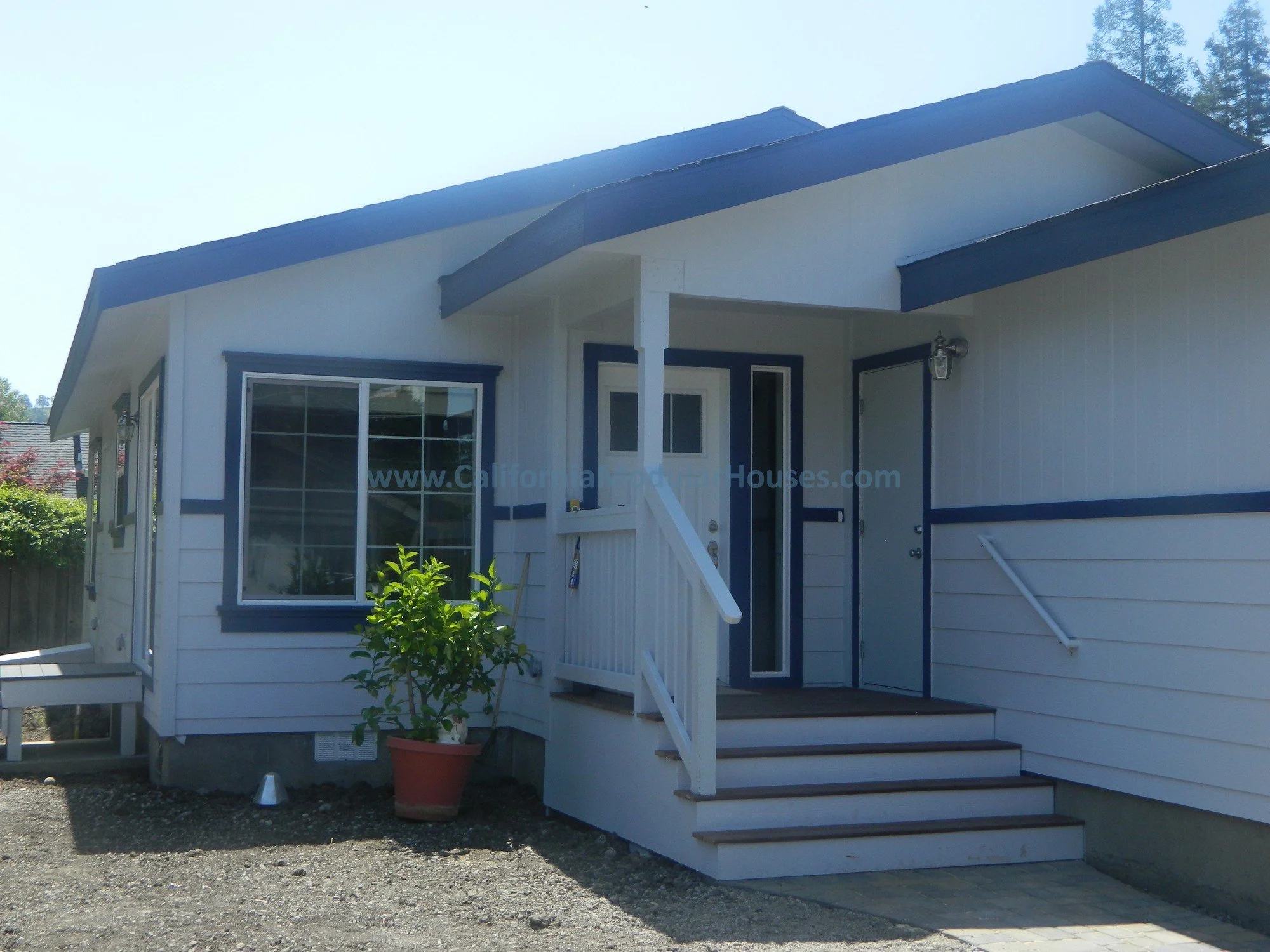 White prefab modular house with blue trim, front steps, potted plant, and a small porch light.