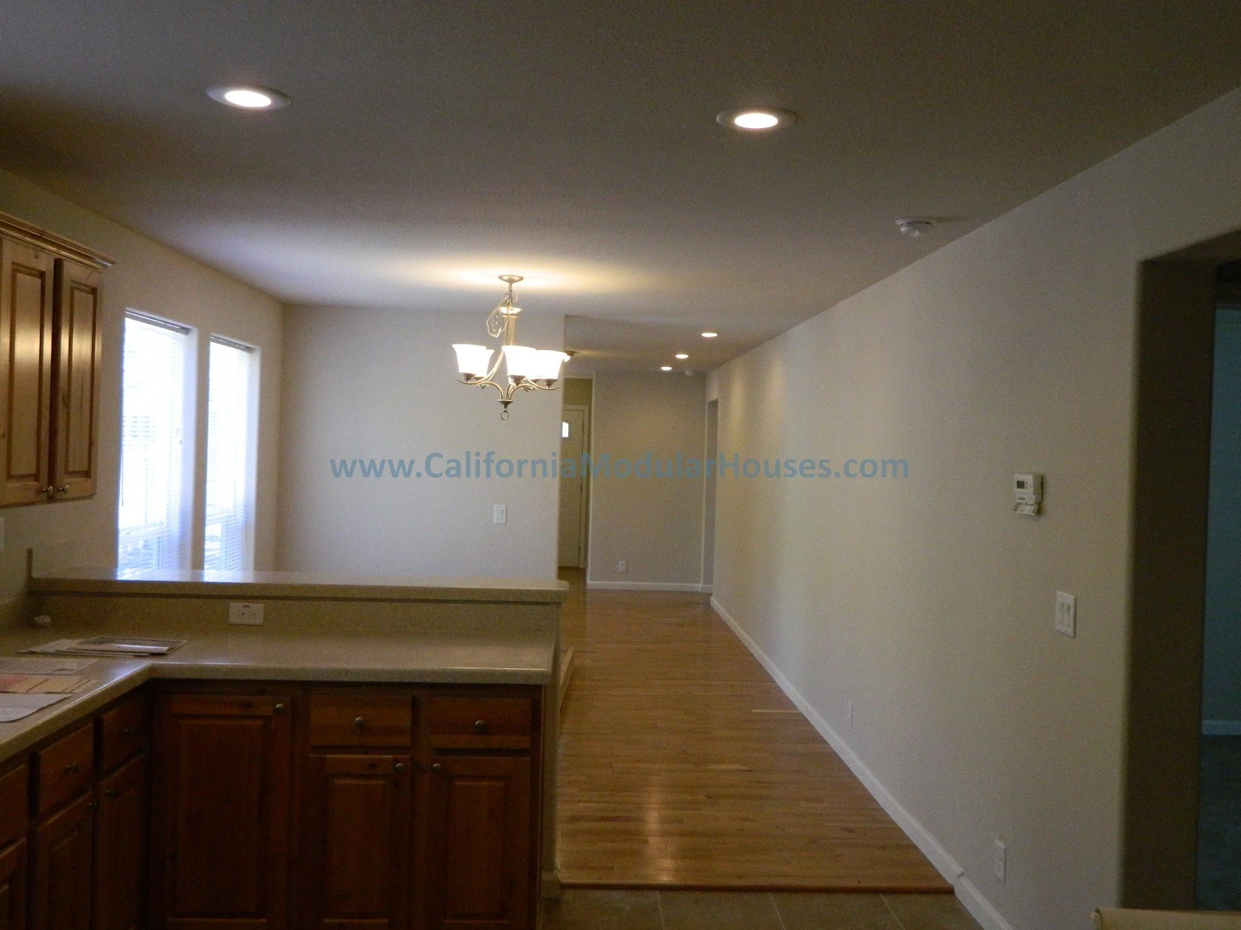 Empty living and dining room with hardwood floors, beige walls, three ceiling lights, a chandelier, and windows with blinds, shown from the kitchen area.