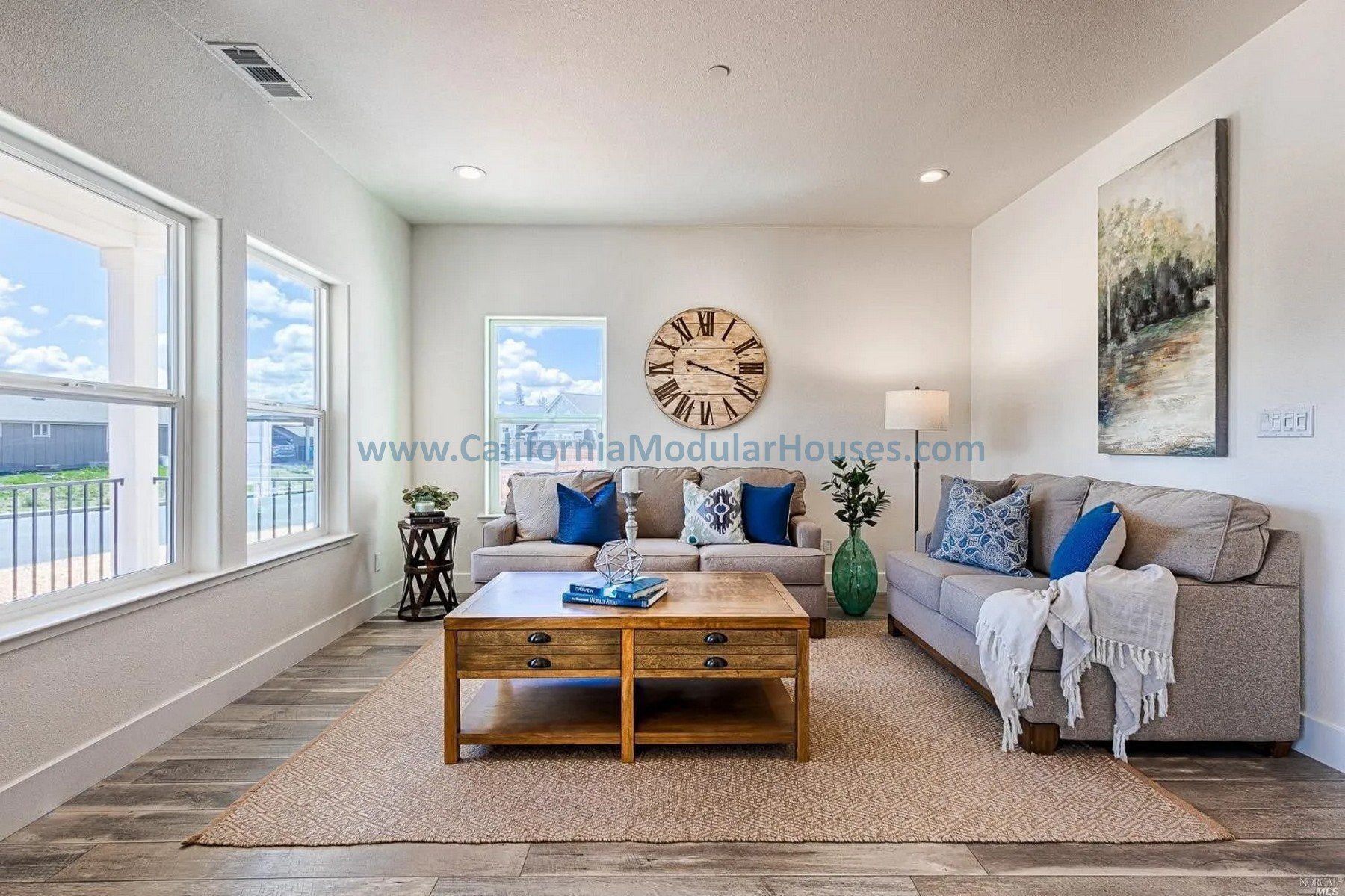 Living room with two beige sofas with blue and patterned pillows, a wooden coffee table, and a beige area rug. There are three windows letting in natural light, a large wall clock, a painting, a floor lamp, and a green vase with a plant.