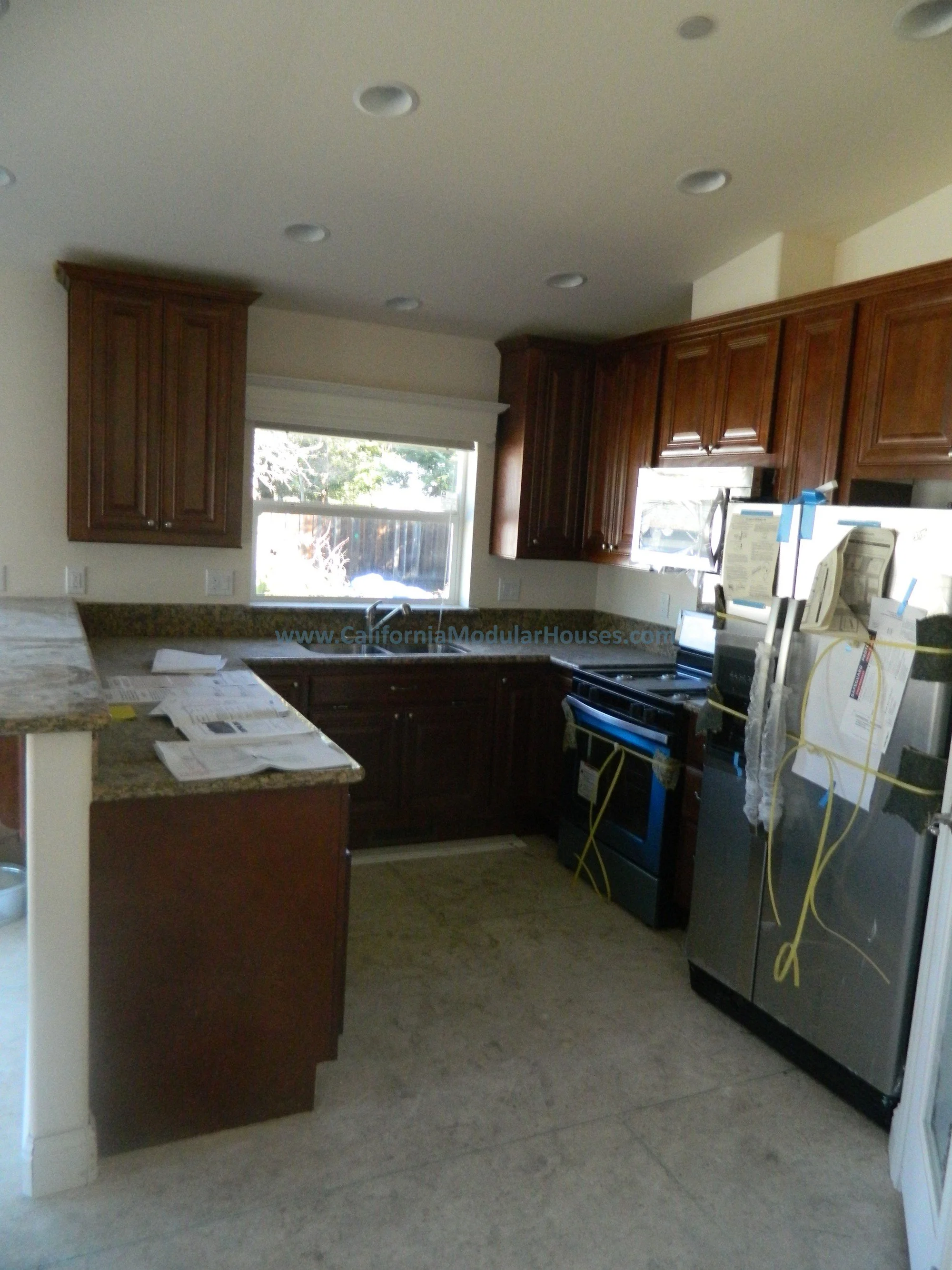 Kitchen with wooden cabinets, a window above the sink, granite countertops, and stainless steel appliances including a refrigerator, stove, and microwave; construction documents and wires attached to appliances indicating ongoing installation.