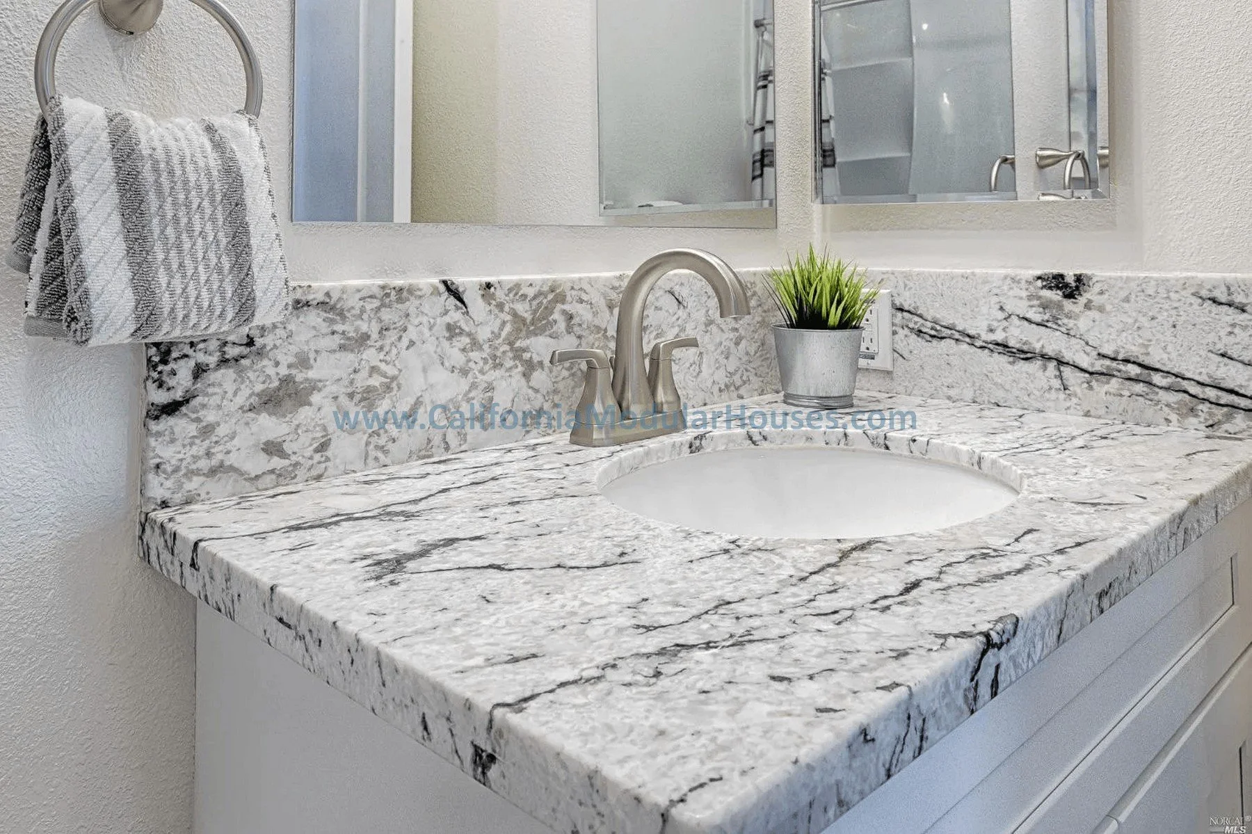 Bathroom vanity with grey and white granite countertop, small potted green plant, silver faucet, and a white towel hanging on a ring.