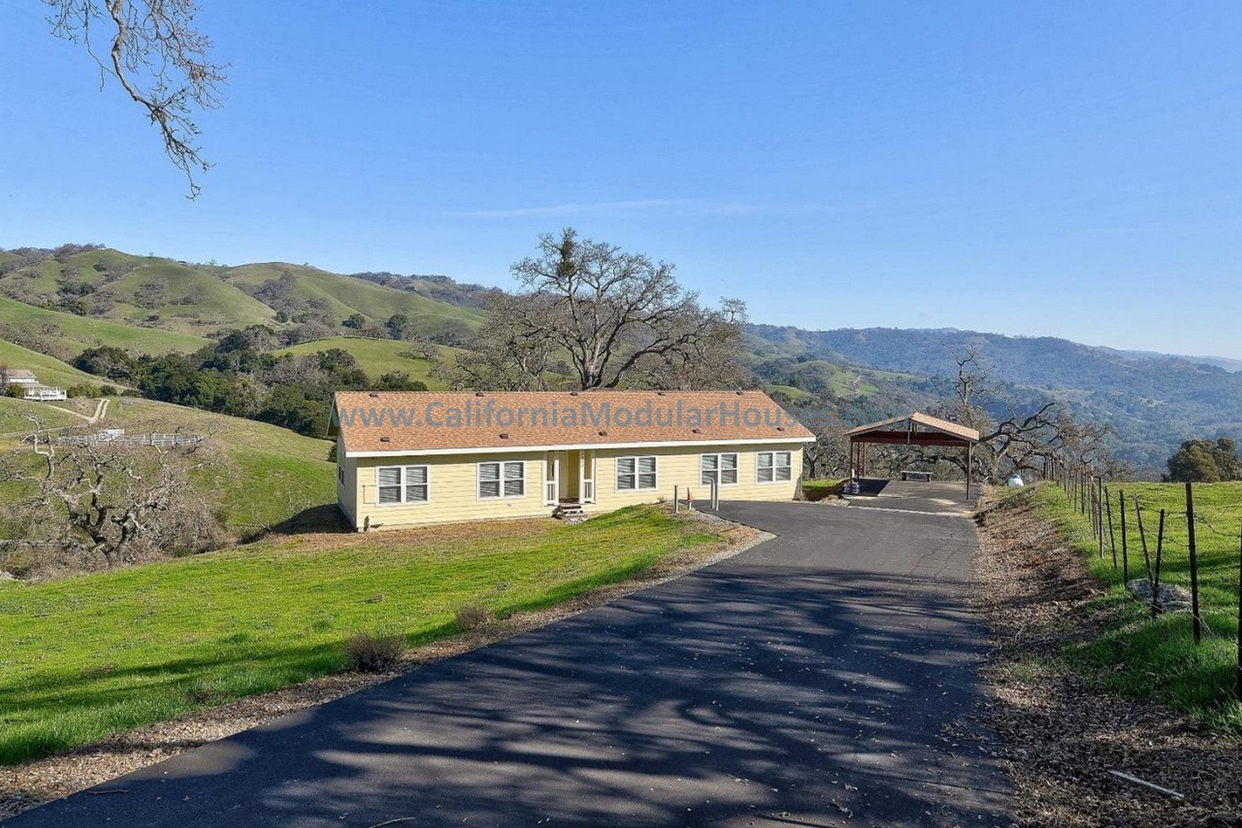 A rural landscape with a yellow house and a covered pavilion on a hillside, with rolling green hills and trees in the background under a clear blue sky.  Bay Area Modular Home, Modular Homes California.