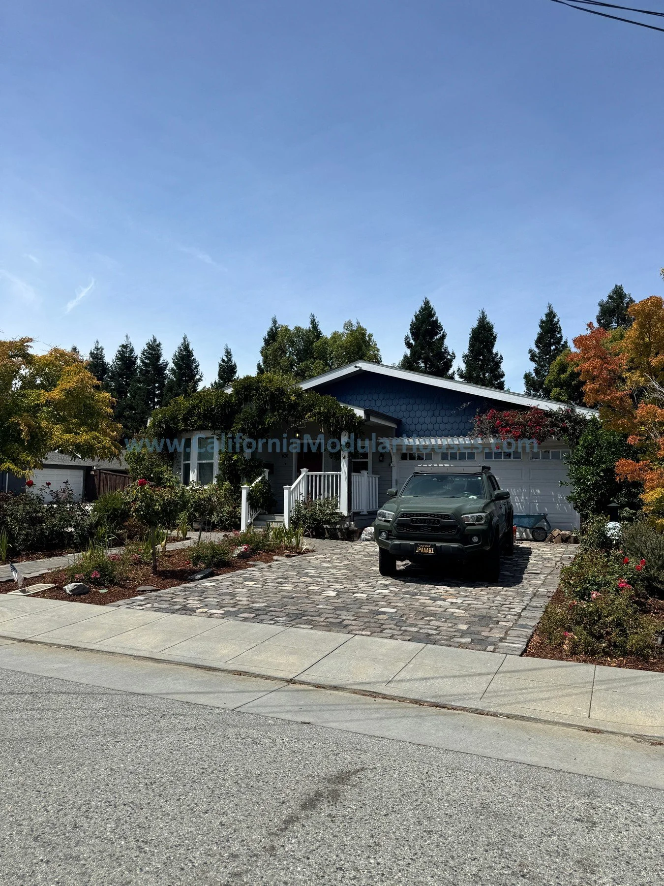 Front yard of a house with a driveway, a parked Toyota vehicle, garden beds, trees, and a clear blue sky.
