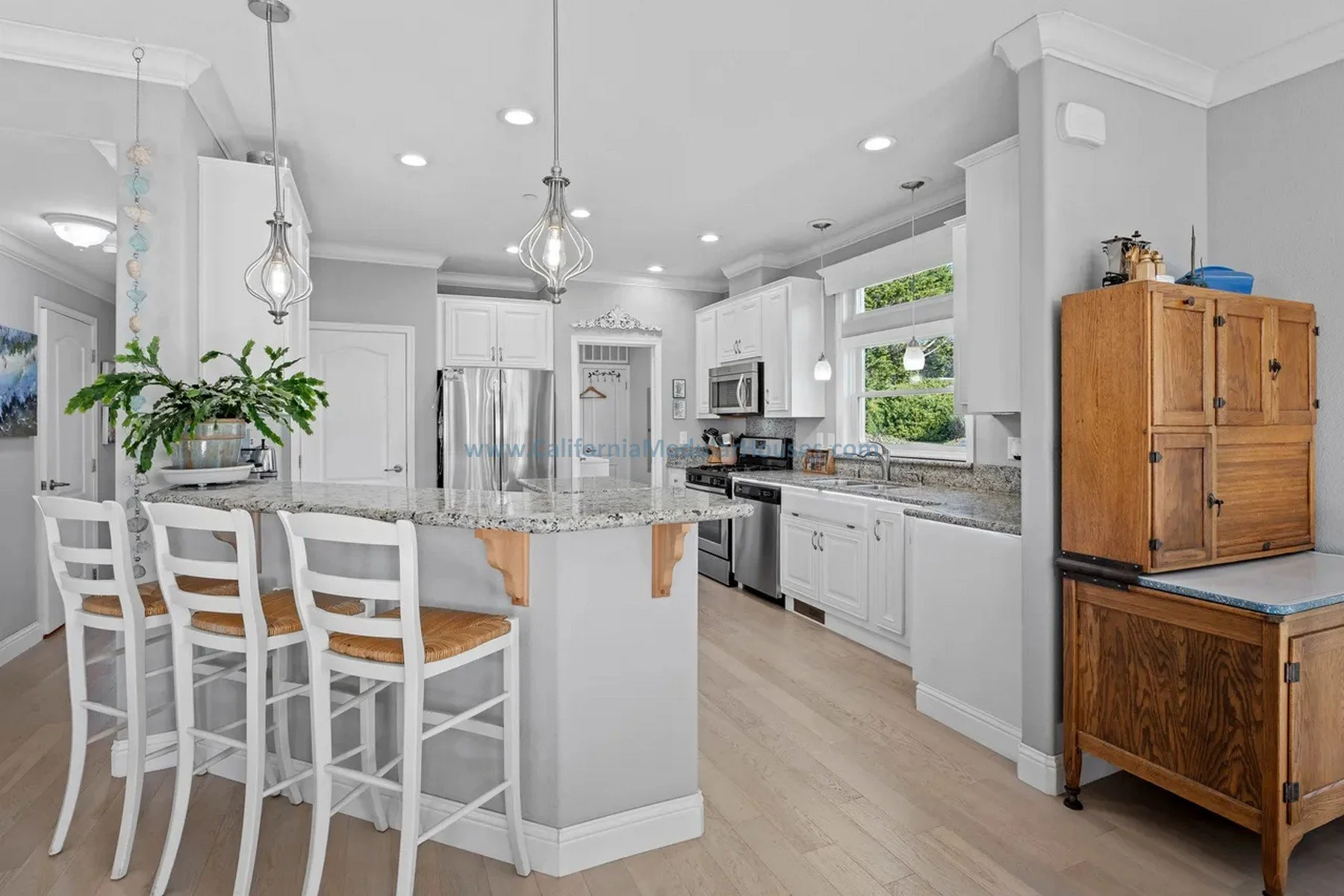 Modern kitchen with white cabinets, granite countertops, stainless steel appliances, a kitchen island with three white chairs, large windows, and a wooden cabinet.  Modular prefab house. California. Cape Cod home with attic finished on-site.