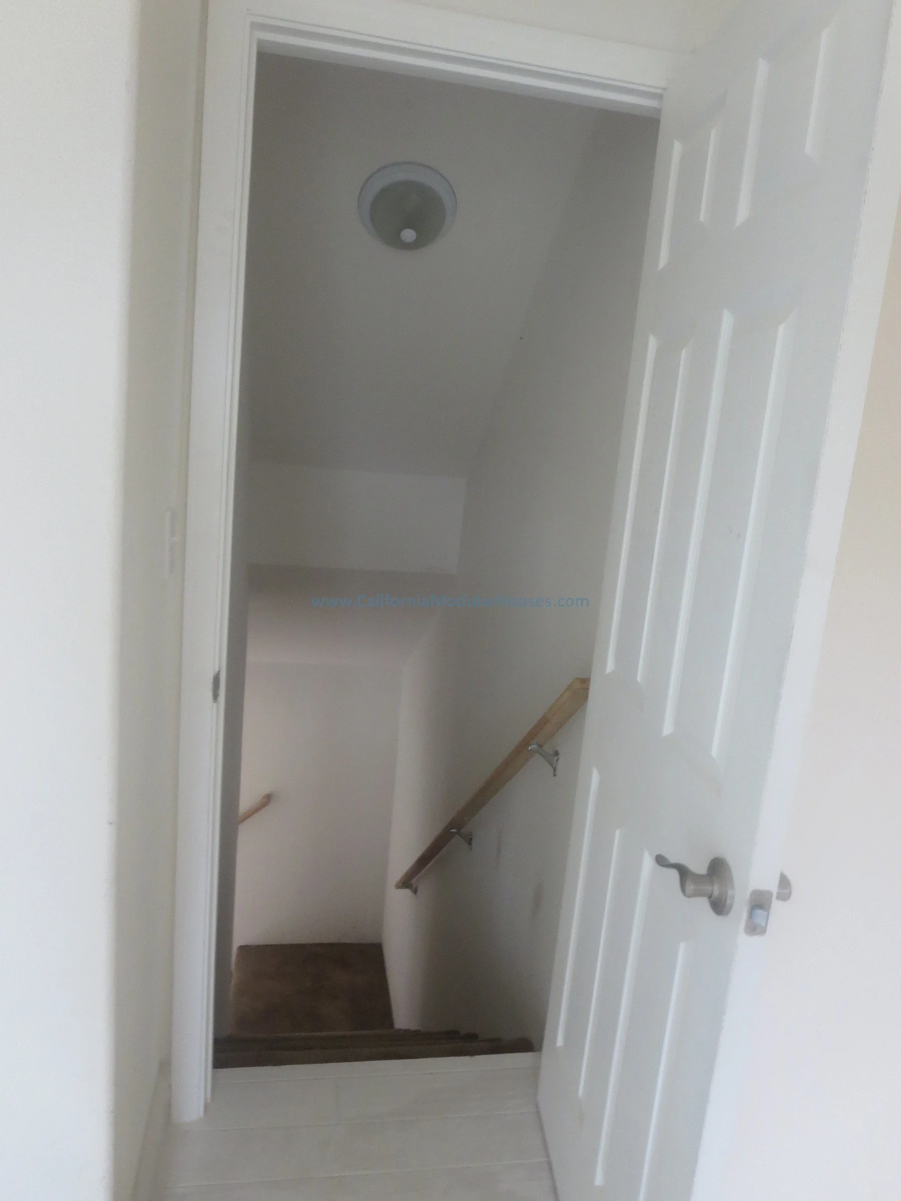 Napa, CA.  California Modular House. California modular home.  Prefab.  Modulars.  Interior view of a staircase with a wooden handrail, white walls, and a closed six-panel door to the right, leading to a lower floor hallway.