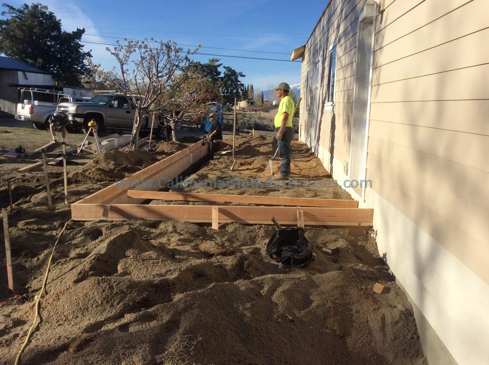 Construction workers building a deck extension at the side of a house with beige siding. Piles of dirt and a partially assembled wooden frame are visible under a blue sky with some clouds.