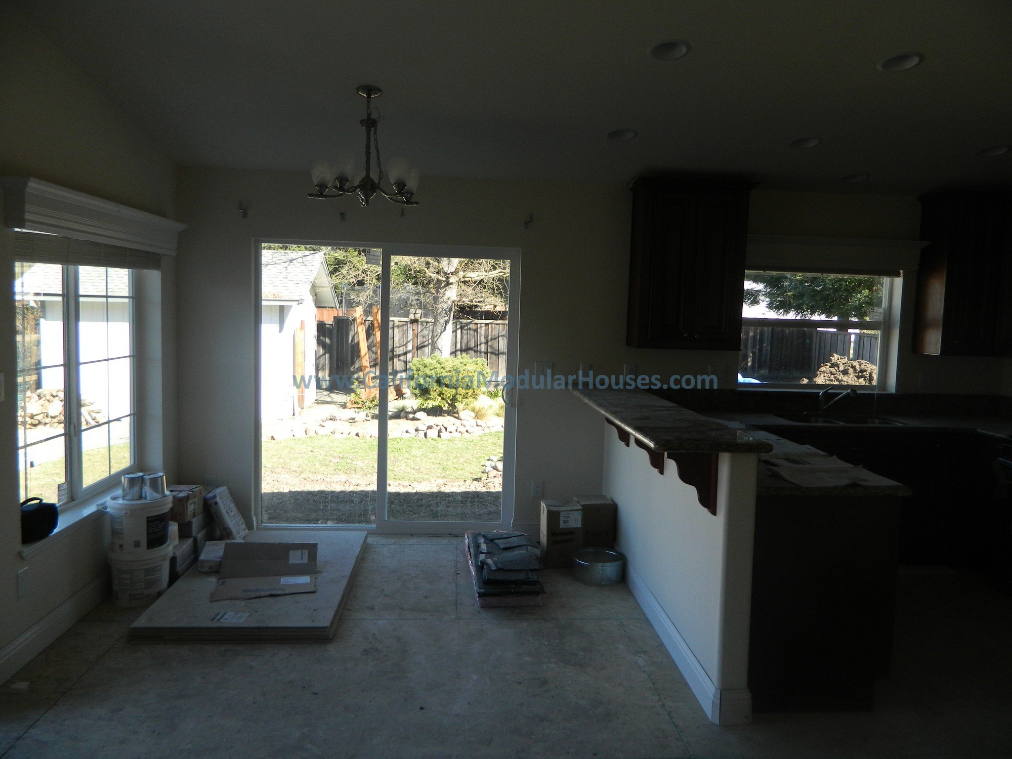 A kitchen and dining area under renovation, with construction supplies on the floor, and a view of a backyard through sliding glass doors.