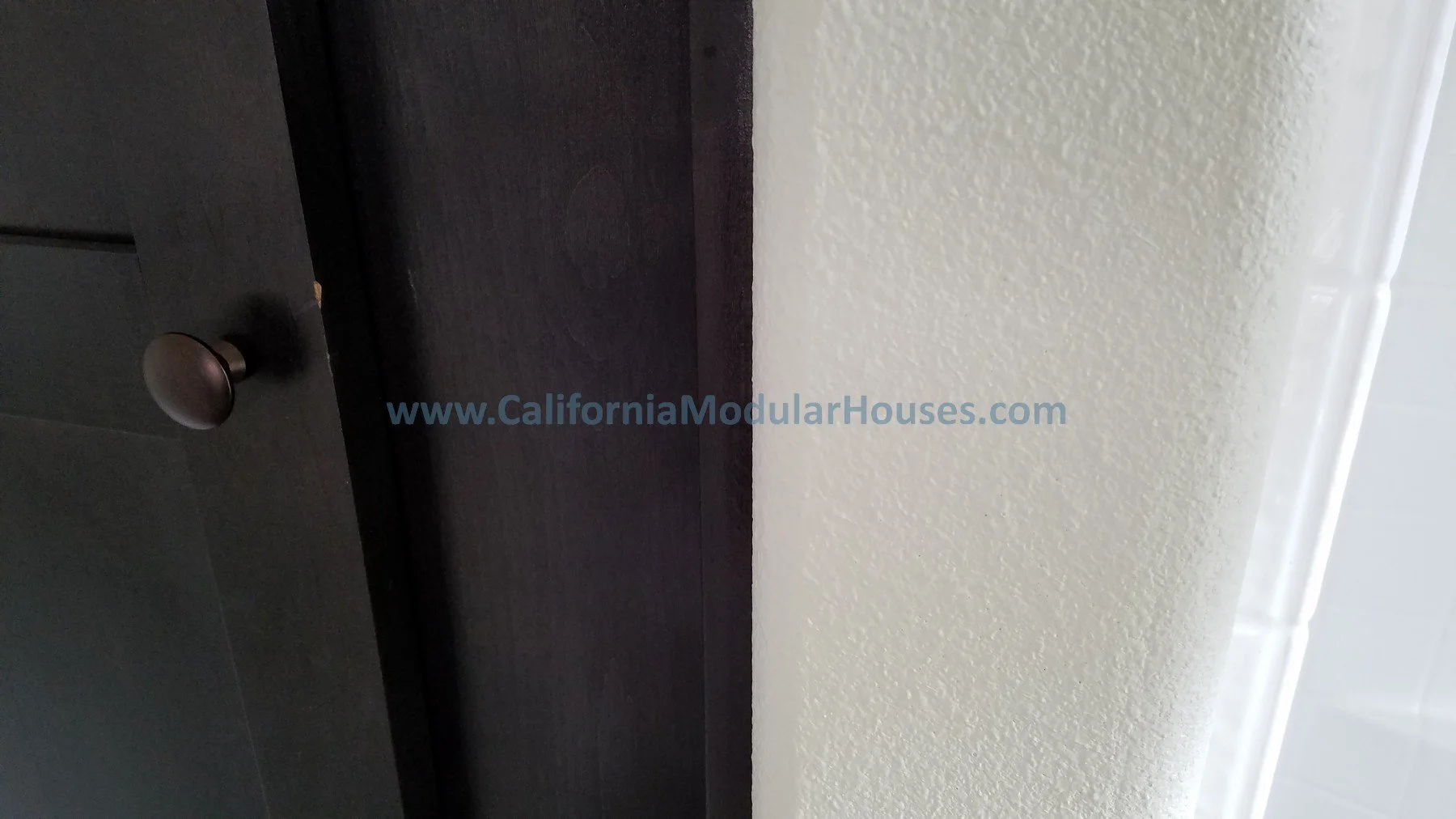 Close-up of a cabinet door with a silver knob, adjacent to a beige textured wall, with a blue watermark reading 'www.CaliforniaModularHouses.com'. California Modular, Prefab Modular Homes California, California Modular Homes,