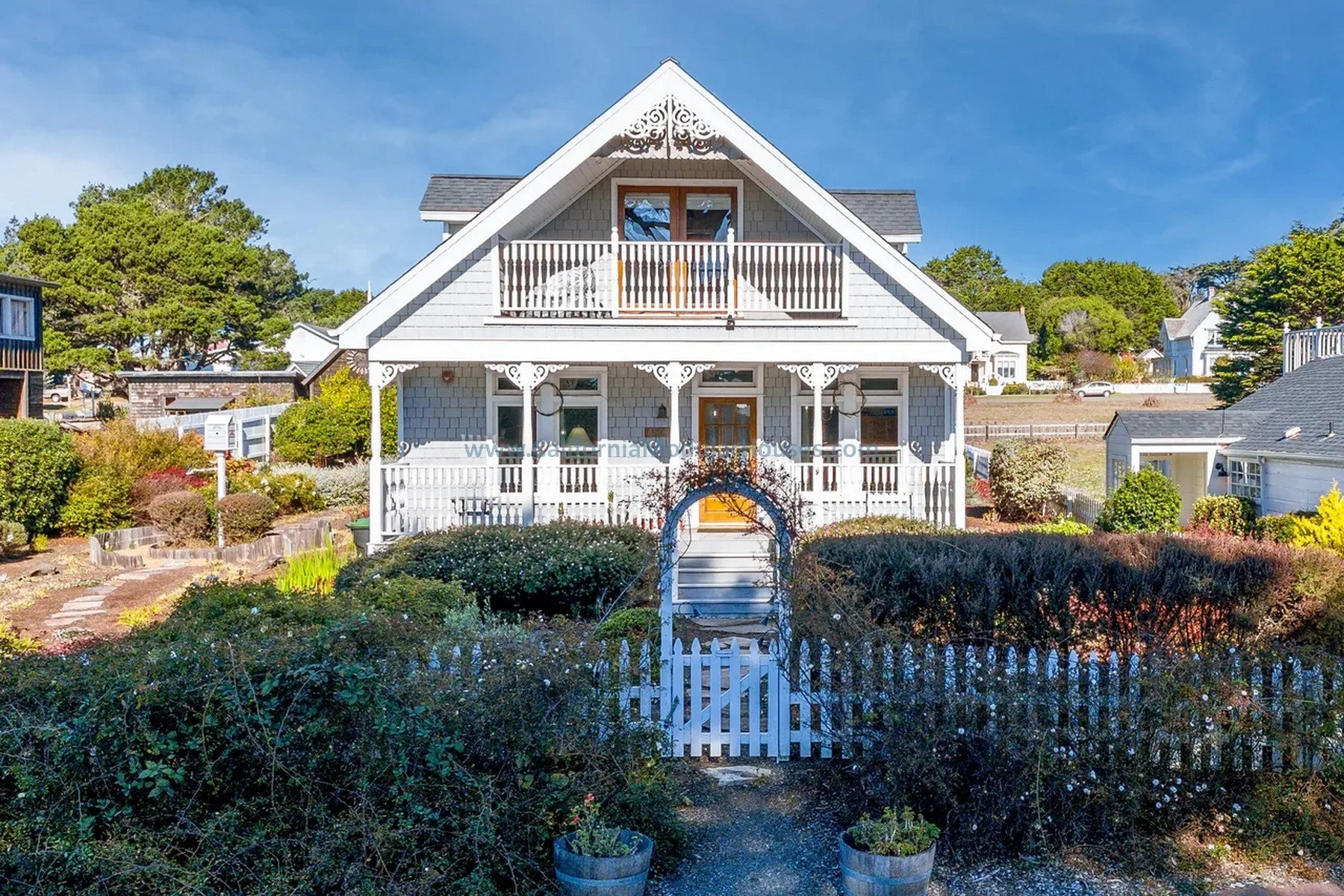 A white Victorian-style house with a steep gabled roof, decorative trim, and a front porch with white railings. The house has a second-floor balcony with wooden doors and a garden in front with bushes, flowers, and a small white picket gate.