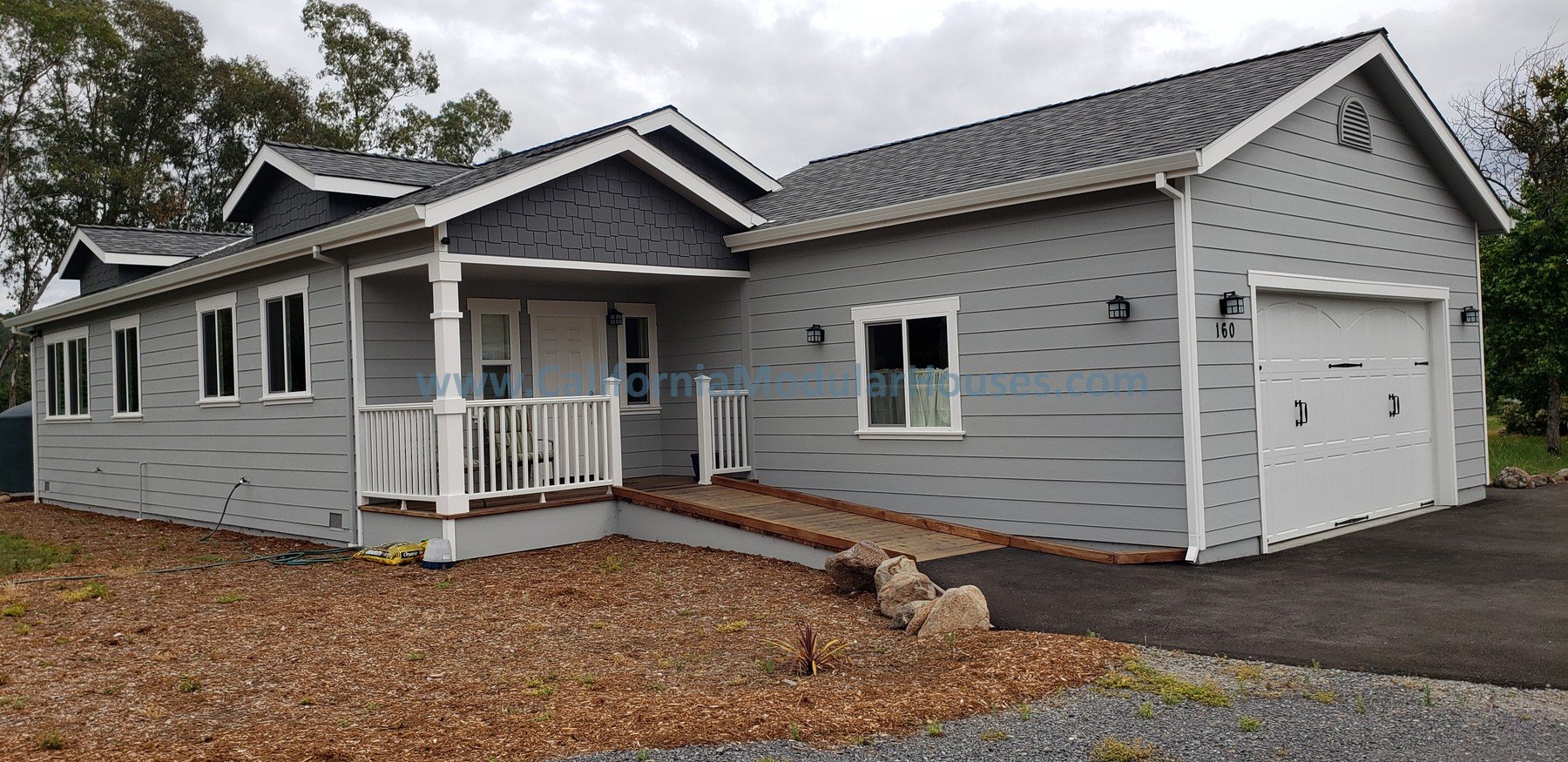 Prefab Modular ADU gray, single-story house with a attached garage, white trim, and a small front porch with a wooden ramp. The house has multiple windows, a gabled roof, and outdoor wall-mounted lights.  