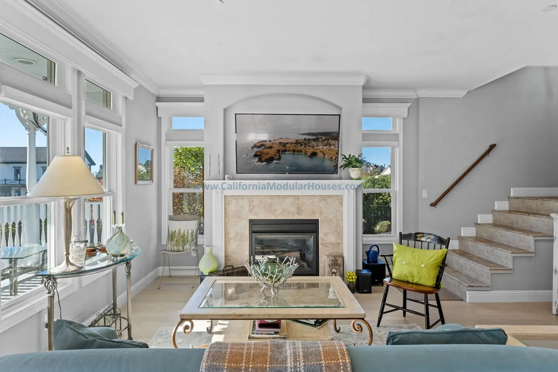 Mendocino, CA. Pre-fab modulars. Custom design coastal commission cape cod modular home. Living room with fireplace, large windows, and TV above the mantel, decorated with vases, a table, and chairs, overlooking a sunny outdoor scene.