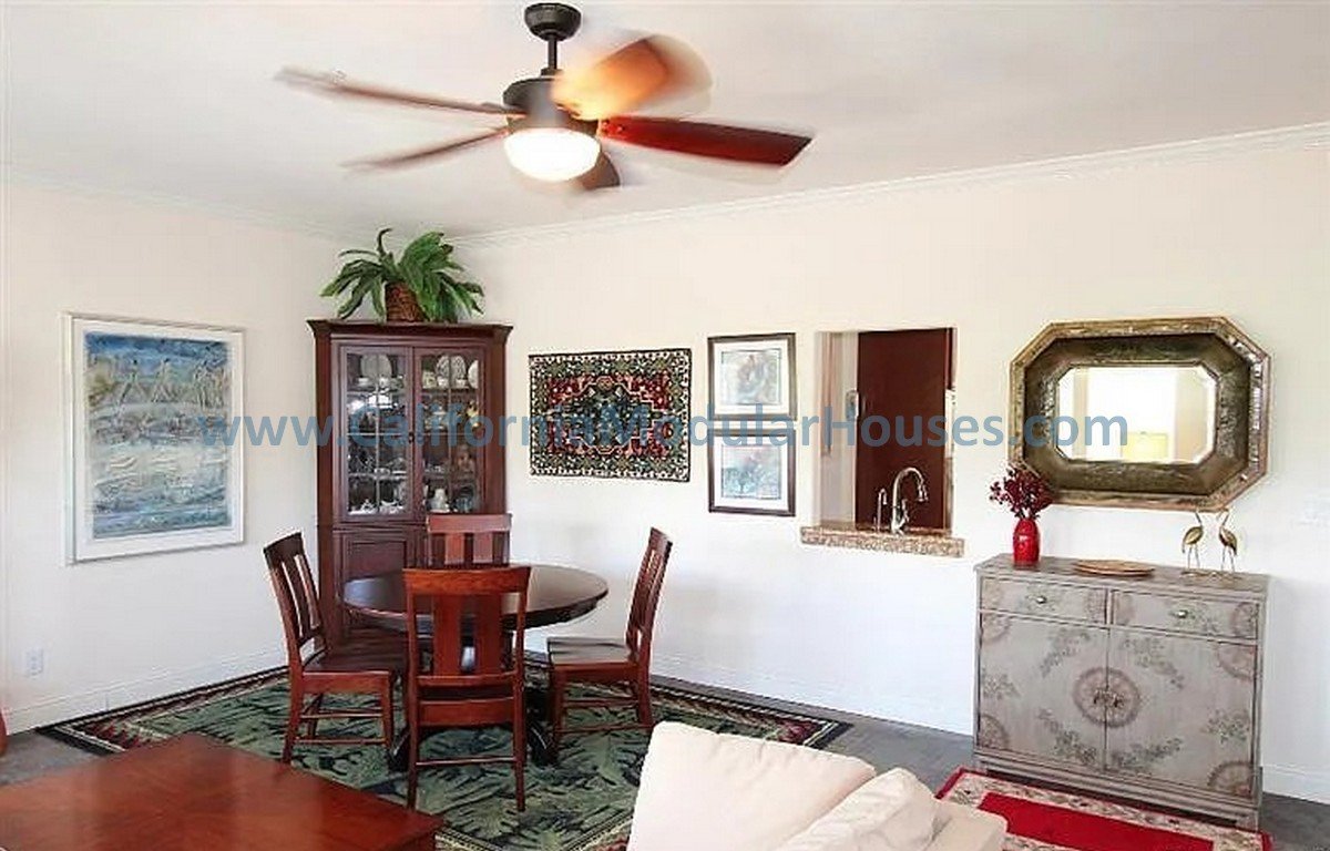 Living room with a ceiling fan, dining table with four chairs, a tall display cabinet, framed artwork, a decorative mirror, a small sideboard, and wall art. There's also a small window opening in the wall.