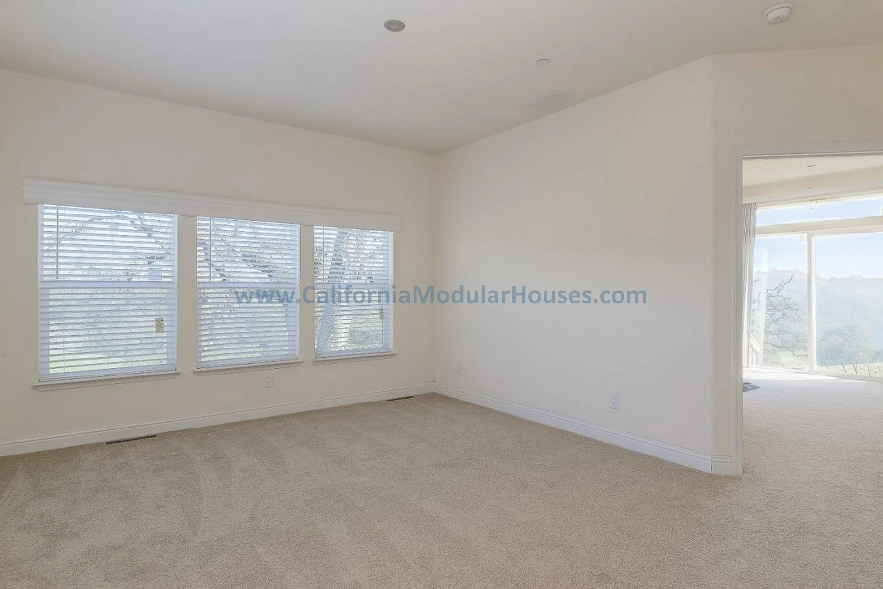 Empty living room with large windows, beige carpet, and white walls.  San Jose Prefab Homes, Modular Homes, Modular Homes California,