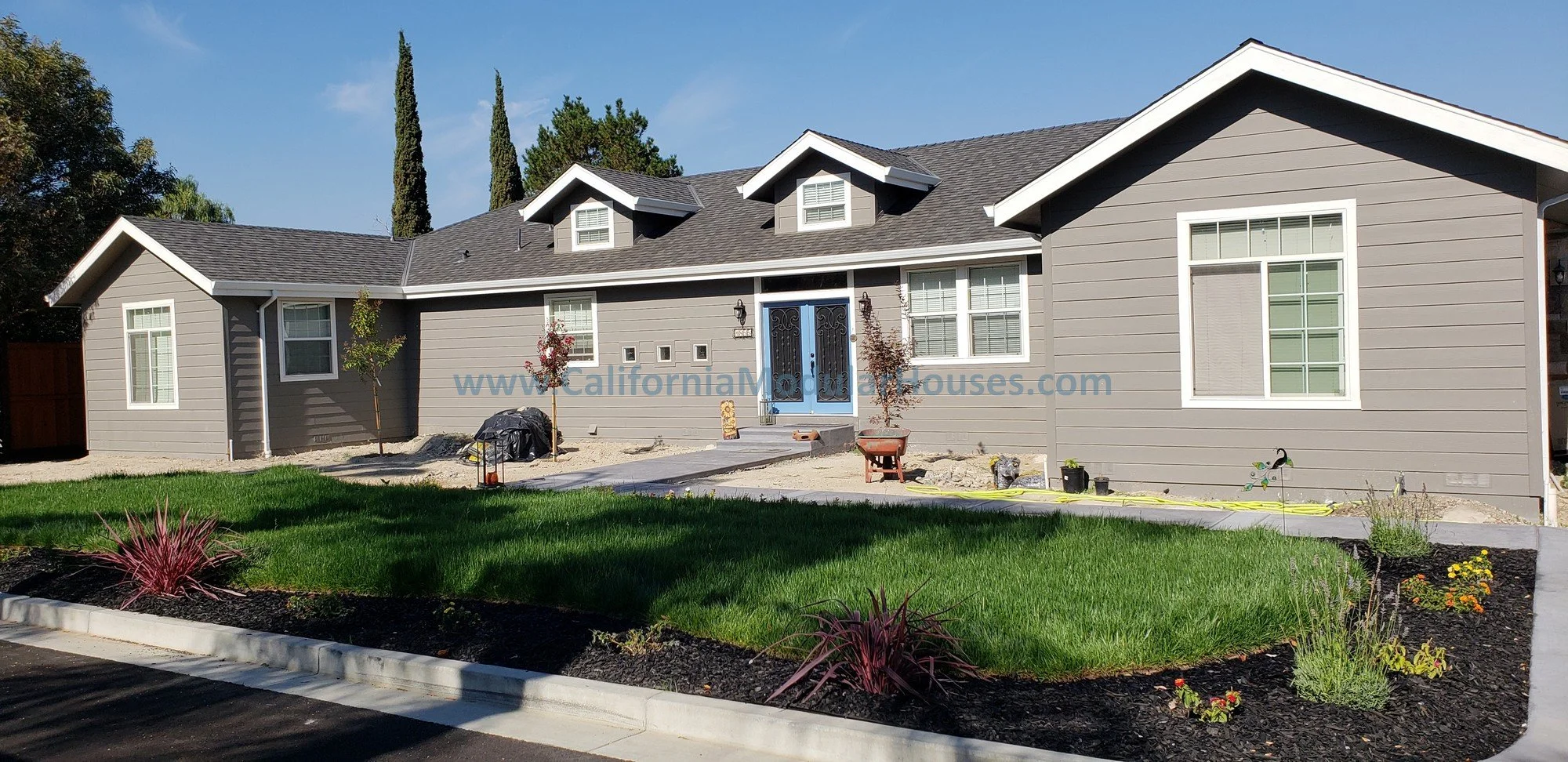 Front view of a newly constructed house with gray siding and white trim, with a small porch and blue front door, surrounded by a green lawn and some landscaped flower beds.