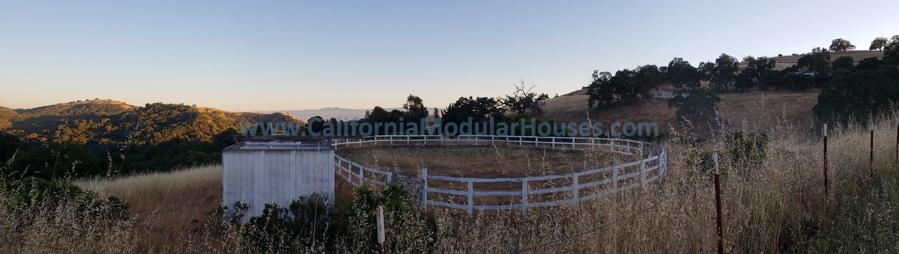 A hilly landscape with dry grass, trees, and a white round fenced area, possibly an arena, with a small white shed in the foreground. The sky is clear with no clouds. Bay Area Modular Home, Prefab Home California,