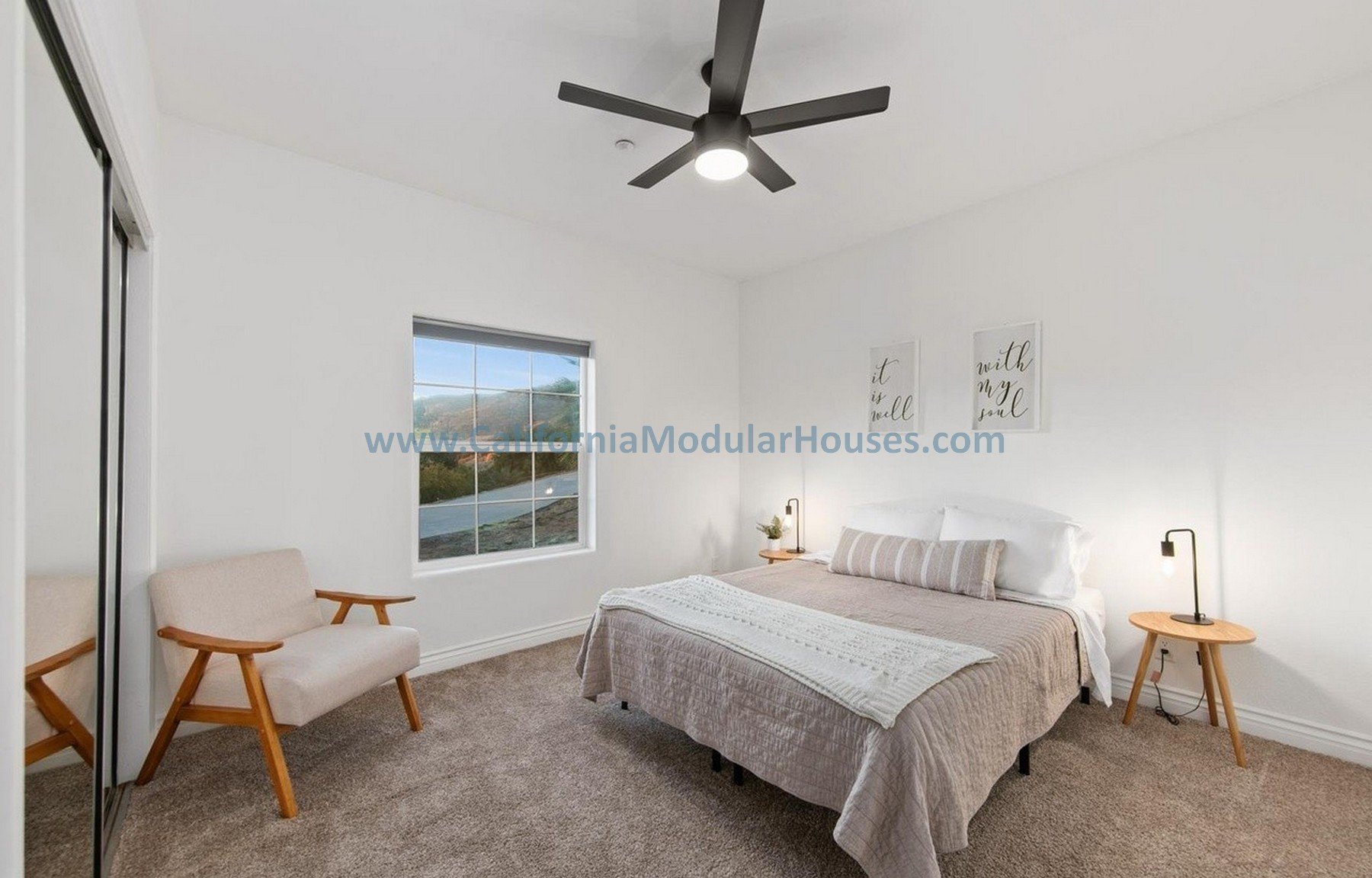 A modern bedroom with white walls, a ceiling fan, a window showing a view outside, a bed with white bedding, and two bedside tables with lamps. There is a beige armchair with wooden arms near a mirrored closet door.