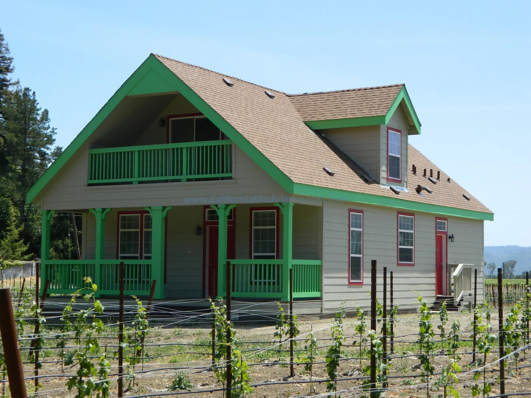 A two-story beige house with green trim and red doors, surrounded by a vineyard with small grapevines supported by trellises, under a clear blue sky.