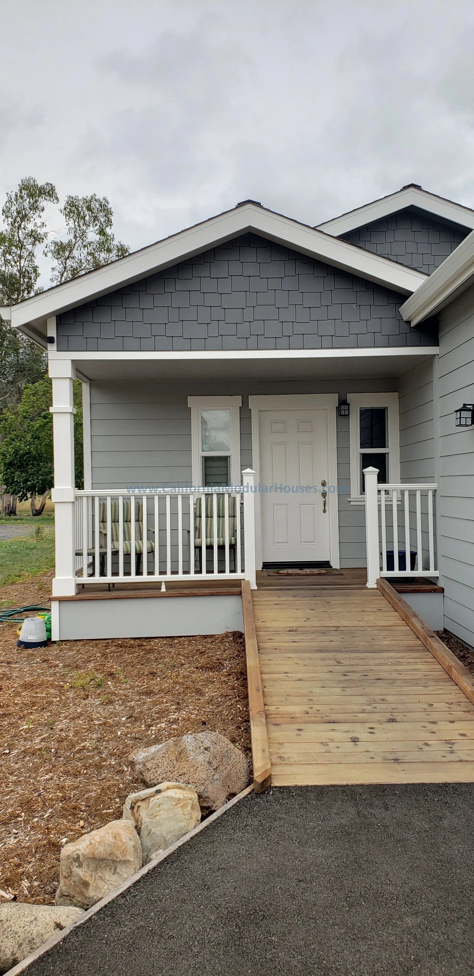 Front view of a new house with a small porch, white railing, and a wooden ramp. The house has gray siding, white trim, and two windows on either side of the white front door. The roof is gray with shingles. Kenwood, Sonoma County, CA. Backyard ADU