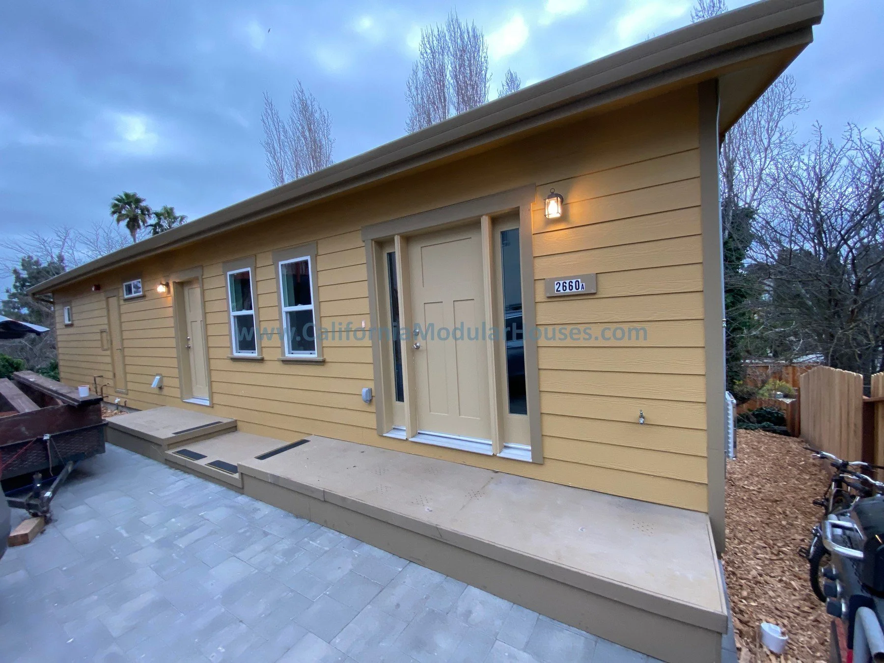 A small, yellow, single-story modular house with three windows and a front door, situated on a tiled patio with steps. Exterior lighting is turned on.  California modular.   Modular CA