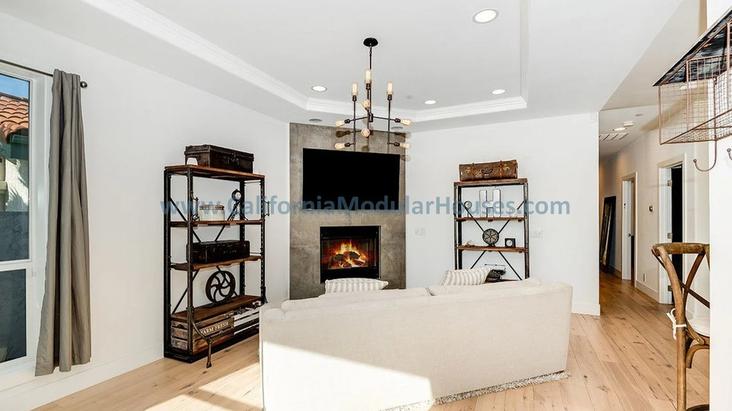 Living room with white walls, a fireplace with a TV above, a beige couch facing the fireplace, a window with gray curtains, and a wooden floor. A modern chandelier hangs from the ceiling.  Modular Home.  Los Angeles, CA.  