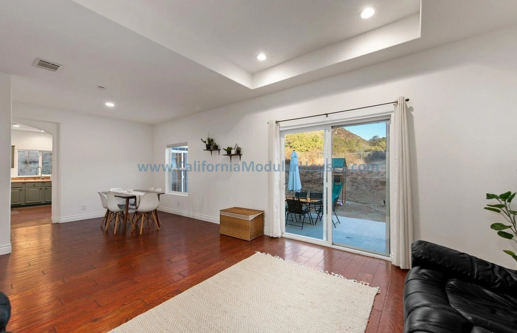 Living room with hardwood floors, white walls, ceiling lights, sliding glass door to patio with outdoor furniture, and a black leather couch.