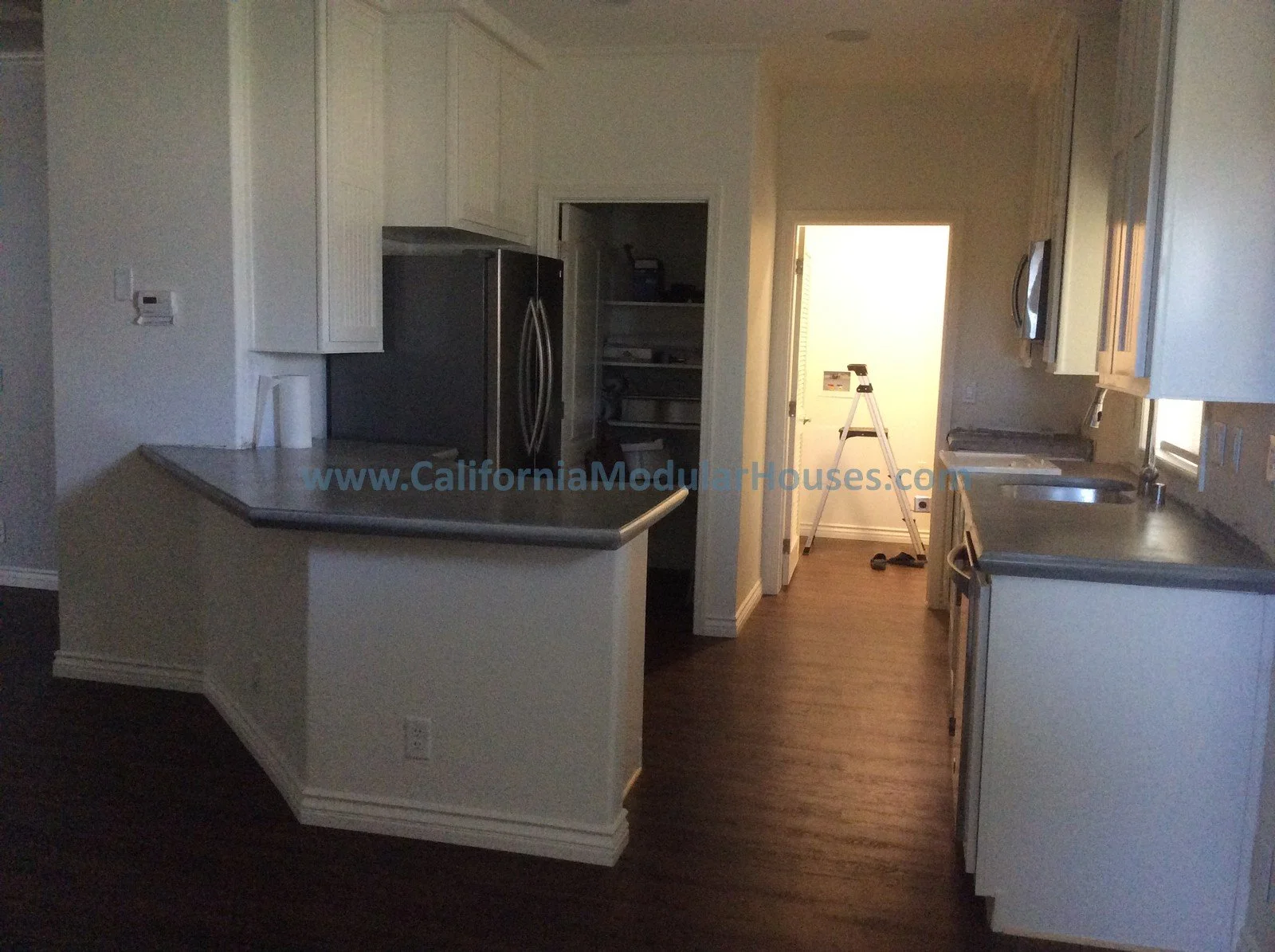 Interior view of a kitchen with white cabinets, a stainless steel refrigerator, wooden flooring, and a doorway leading to a laundry area with a step ladder.