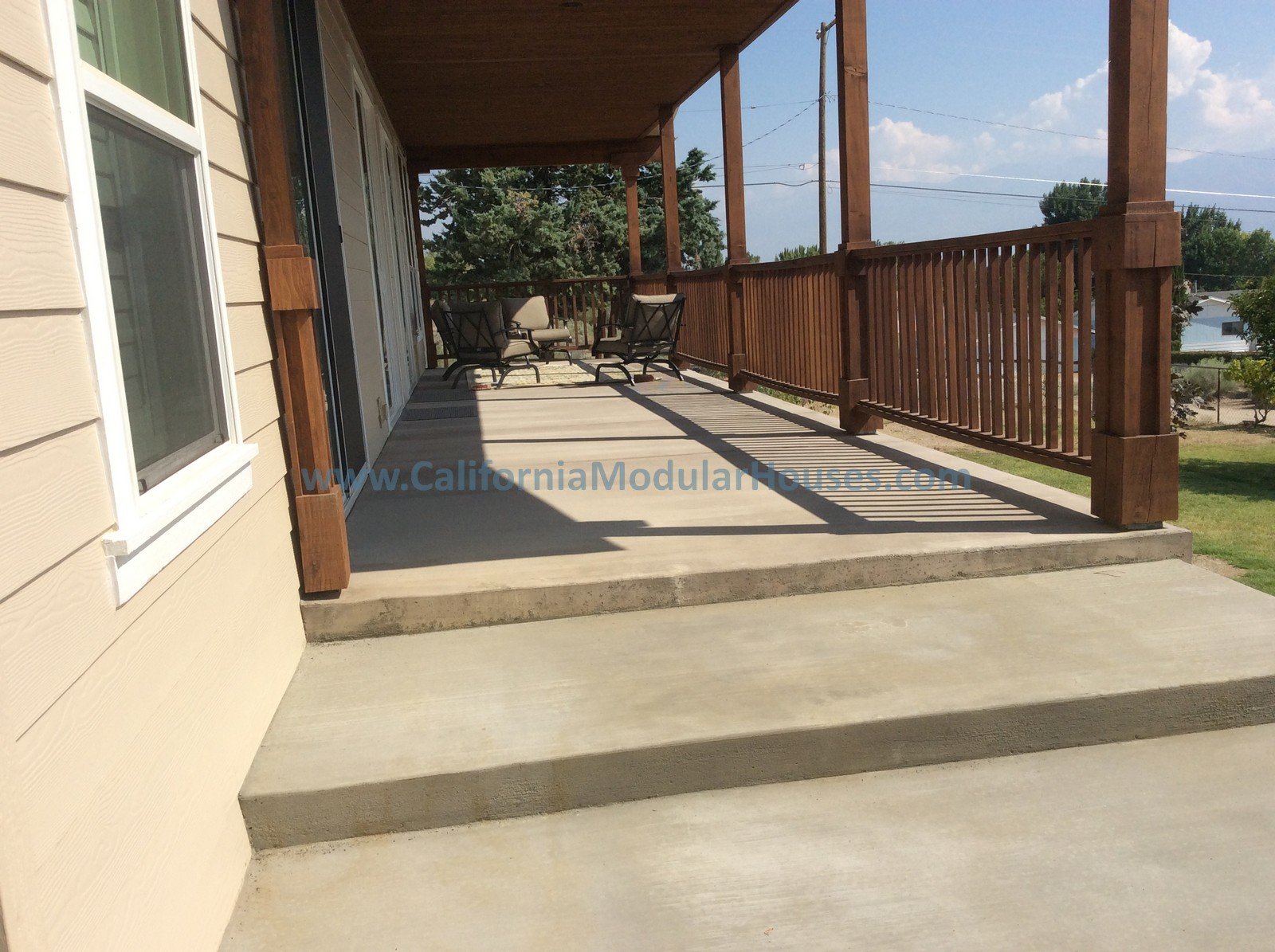 A large wooden balcony attached to a beige house with a sliding glass door, featuring a concrete floor, wooden railing, and outdoor furniture including chairs and a small table.
