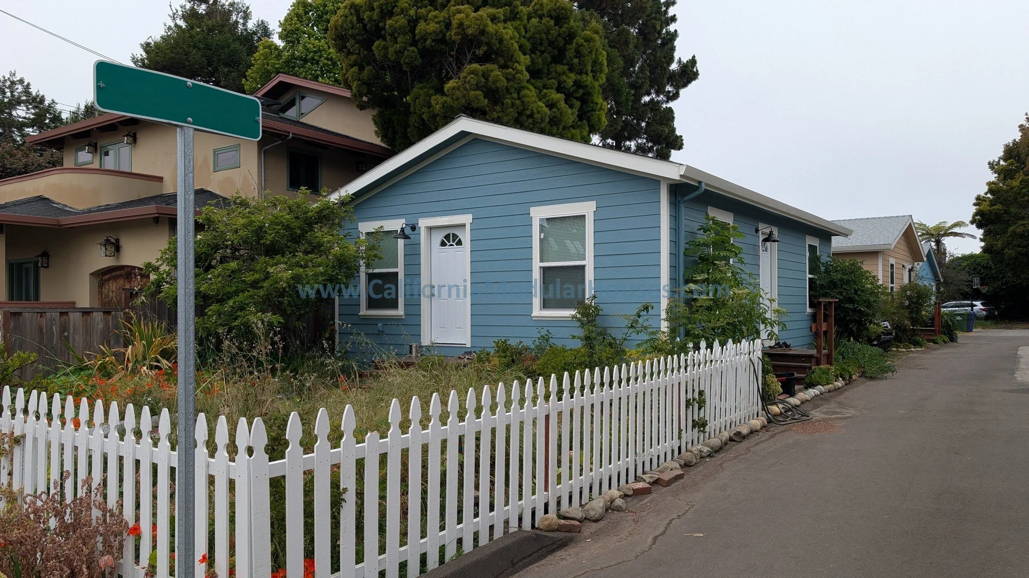 A blue house with a white picket fence along a street. The house has white trim and a small front porch. Surrounding garden with bushes and small trees. Nearby houses are visible in the background, and a large tree extends over the roof. An empty str