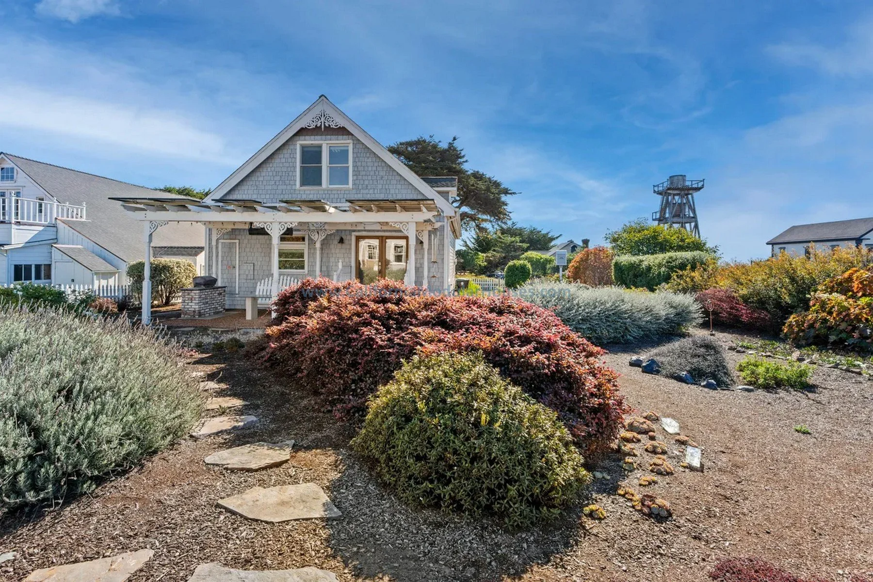 Mendocino, CA. Pre-fab modulars. Custom design coastal commission cape cod modular home.  A quaint white house with a front porch surrounded by colorful bushes and a well-maintained garden under a blue sky.