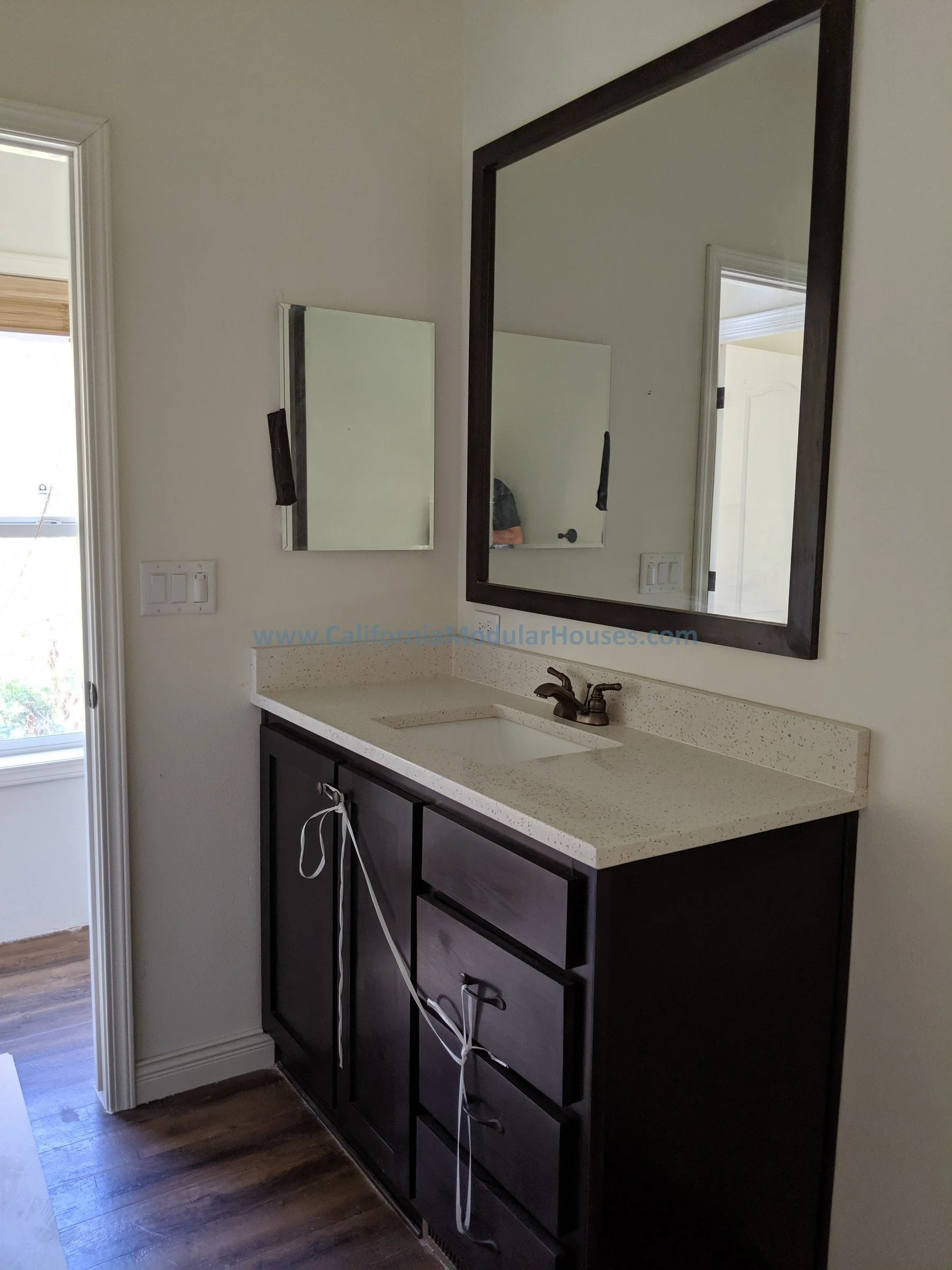 Bathroom vanity with a dark wooden cabinet, a beige speckled countertop, a metallic sink faucet, a large framed mirror, a smaller mirror, and multiple cords hanging from the cabinet handle. Modular House California.