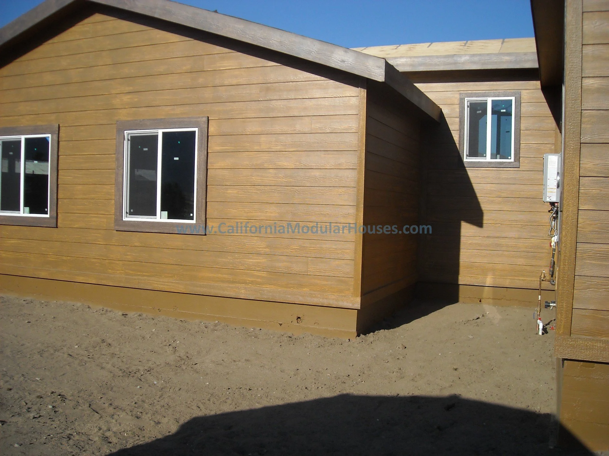 Side view of a newly constructed modular house with wooden siding and three windows, set on a dirt yard under a clear blue sky.  Walker Basin, Kern County, California.  