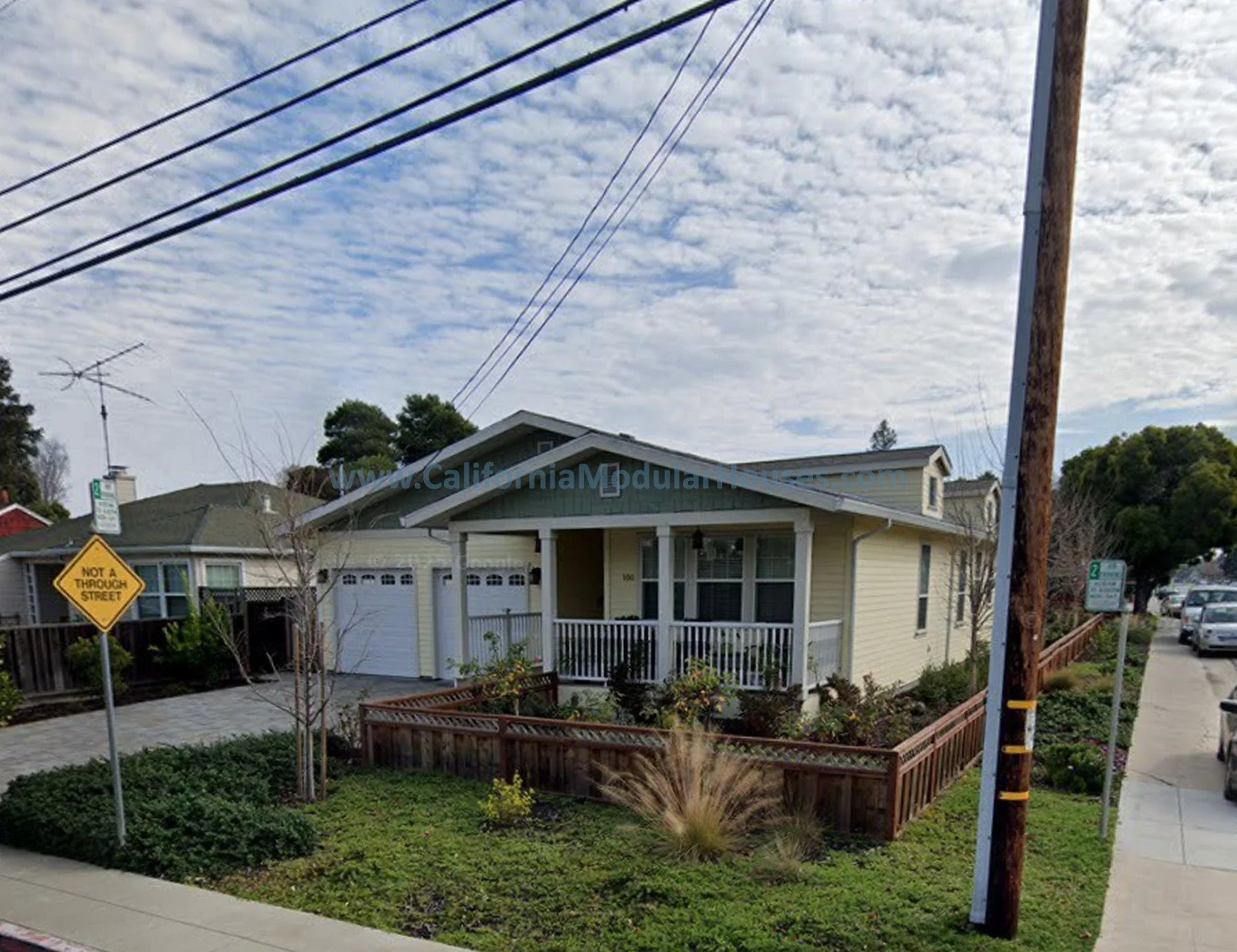 A yellow house with a front porch, surrounded by a small garden with shrubs and plants, in a suburban neighborhood. The sky above is partly cloudy with visible power lines.  San Mateo City, San Mateo County, CA. Pre-Fabricated Homes,
