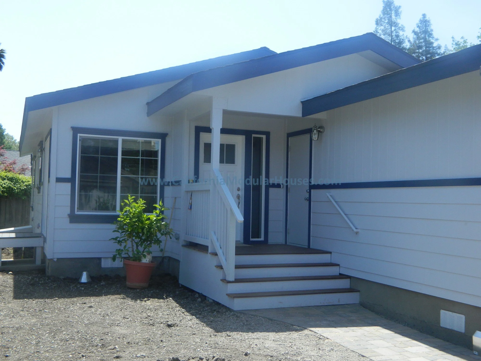 Exterior view of a white house with blue trim, small front porch with stairs, window with grid pattern, potted plant on porch, and a gravel walkway.