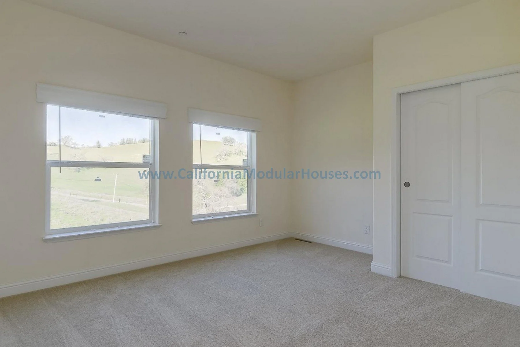 Empty bedroom with two windows overlooking green hills, white walls, beige carpet, and a white closet door. Bay Area Prefab Homes, Prefab Modular Homes California, California Modular,