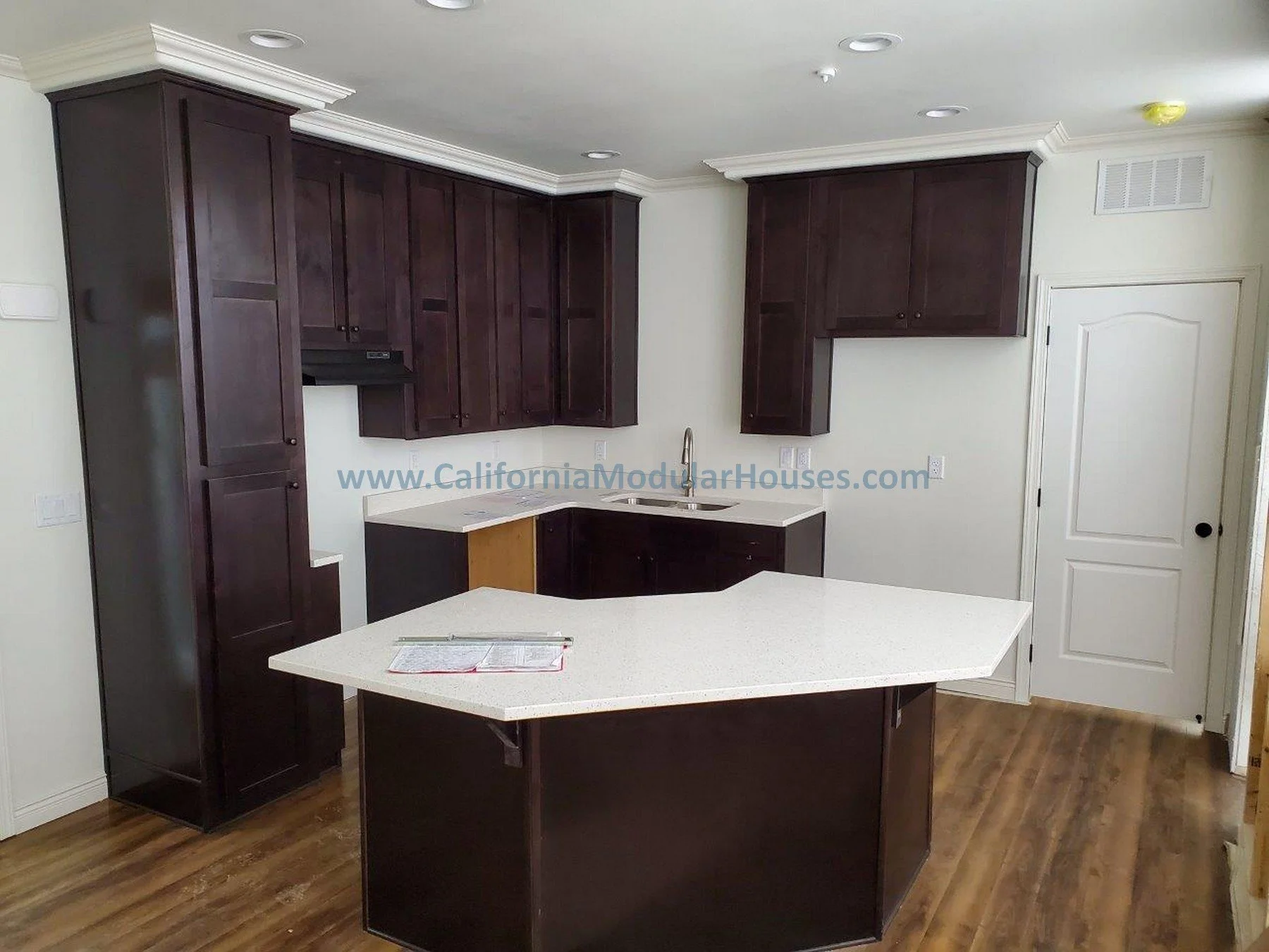 Kitchen with dark wood cabinets, white countertops, and hardwood floors. There is a kitchen island with a book and pen on top, a sink under the cabinets, and a white door in the corner.  Prefab Home California, Modular Homes in California, Modular.  