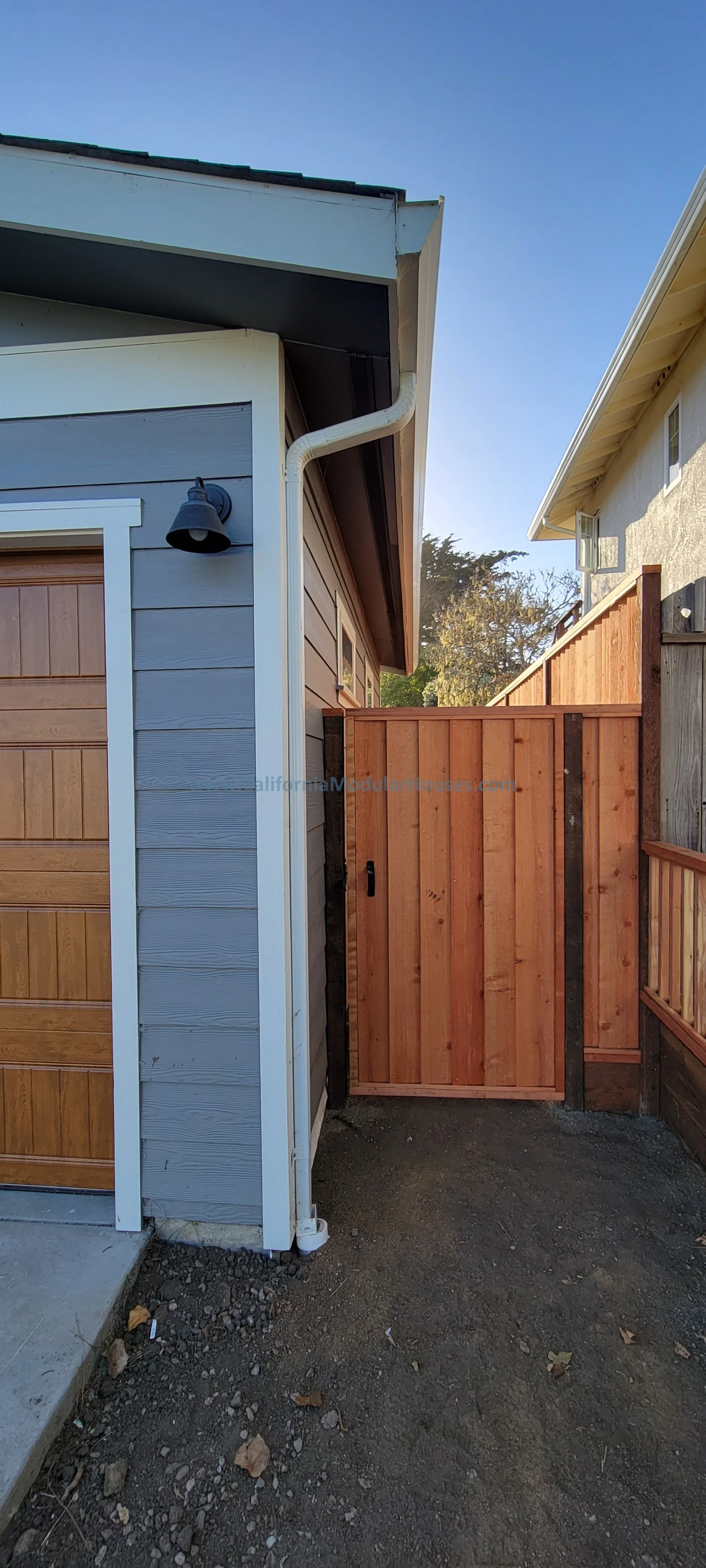 Side view of a residential backyard with a wooden privacy fence, a gray house with a garage door, a black outdoor wall light, and a small gate in the fence, with trees and a clear blue sky in the background.