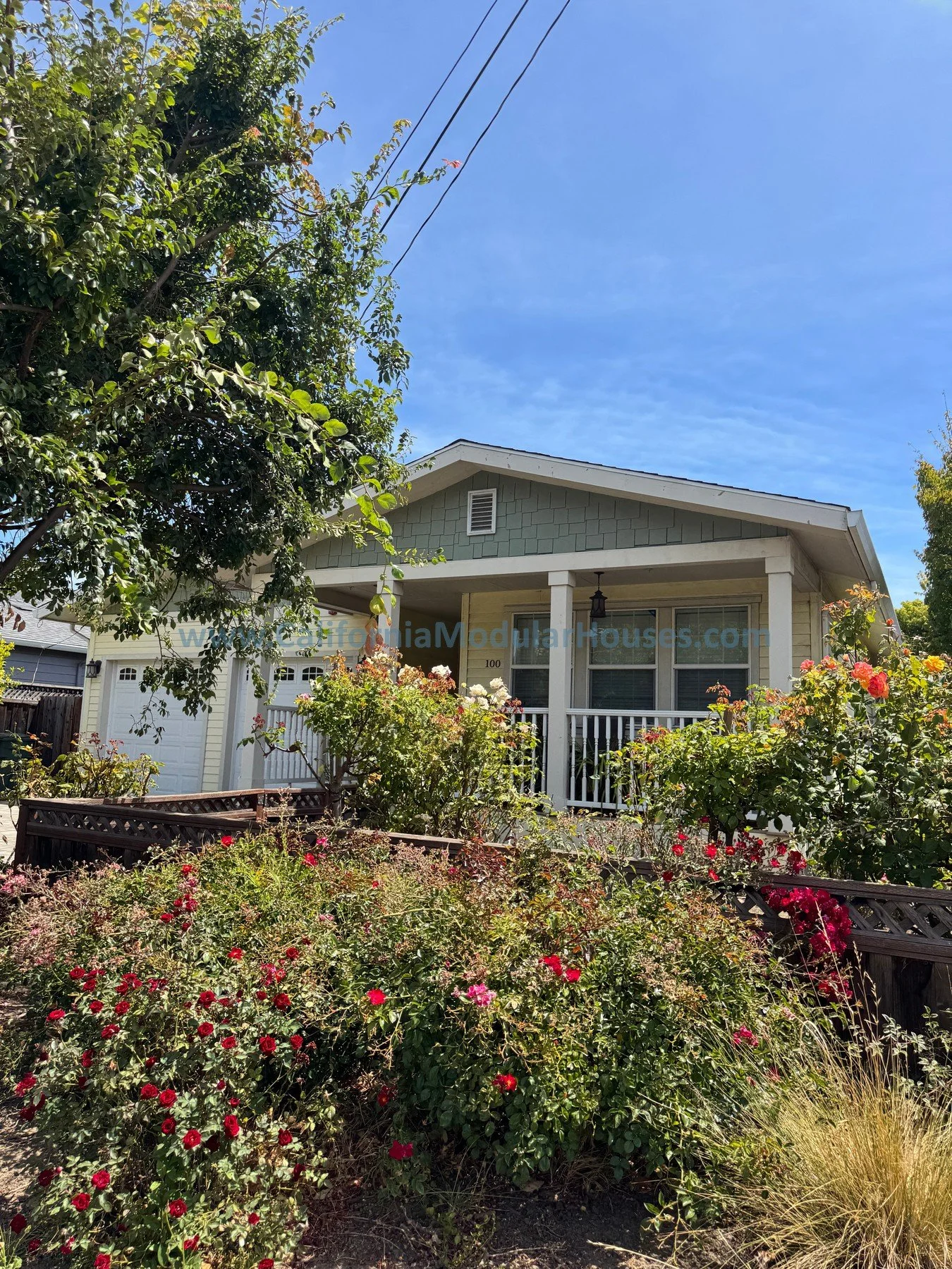 A cozy house with a small front porch, surrounded by colorful flowers and lush green trees on a sunny day with a blue sky.