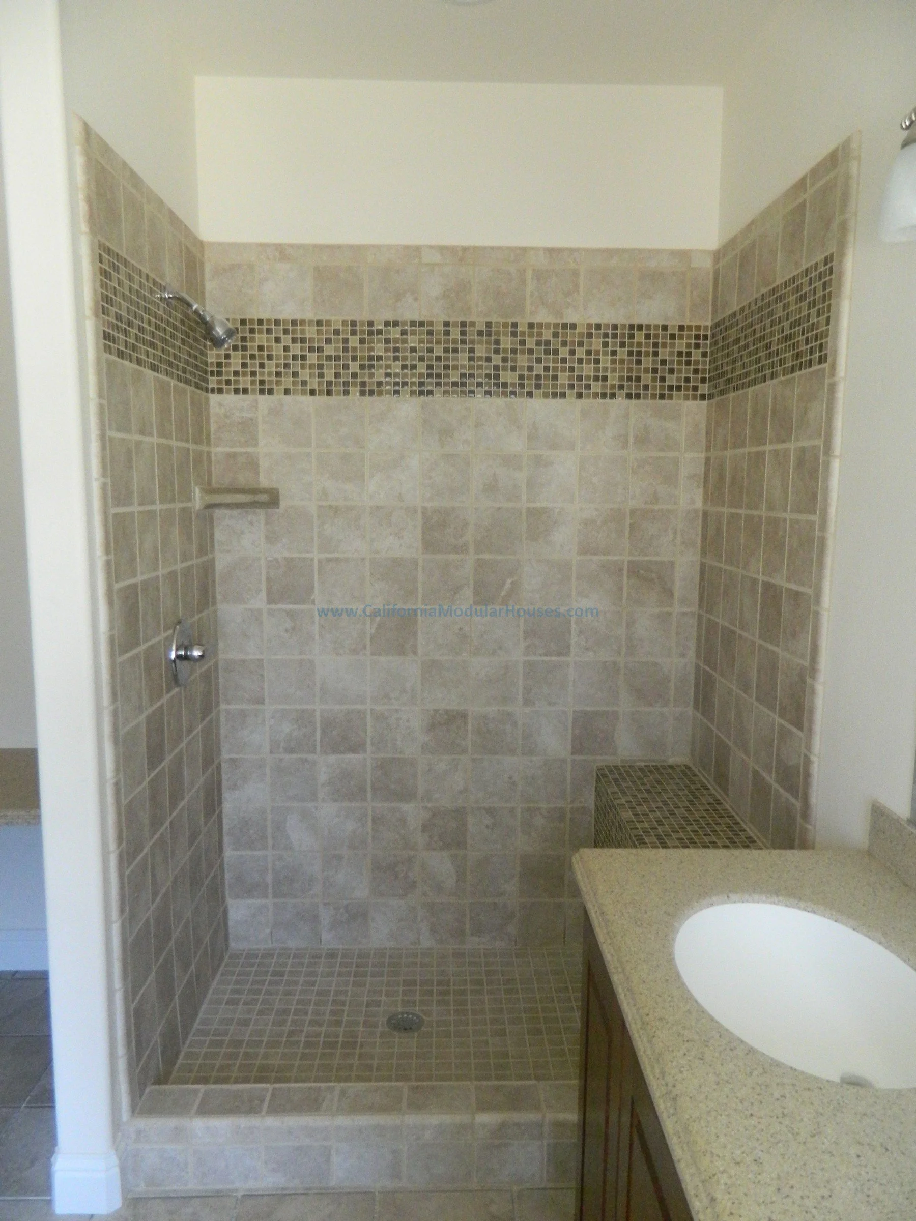 Bathroom shower area with beige and brown tiled walls, built-in shelf, a showerhead, and drain on the tiled floor, adjacent to a bathroom vanity with a granite countertop and an integrated sink. City of San Mateo, San Mateo County, CA.  Modular home.