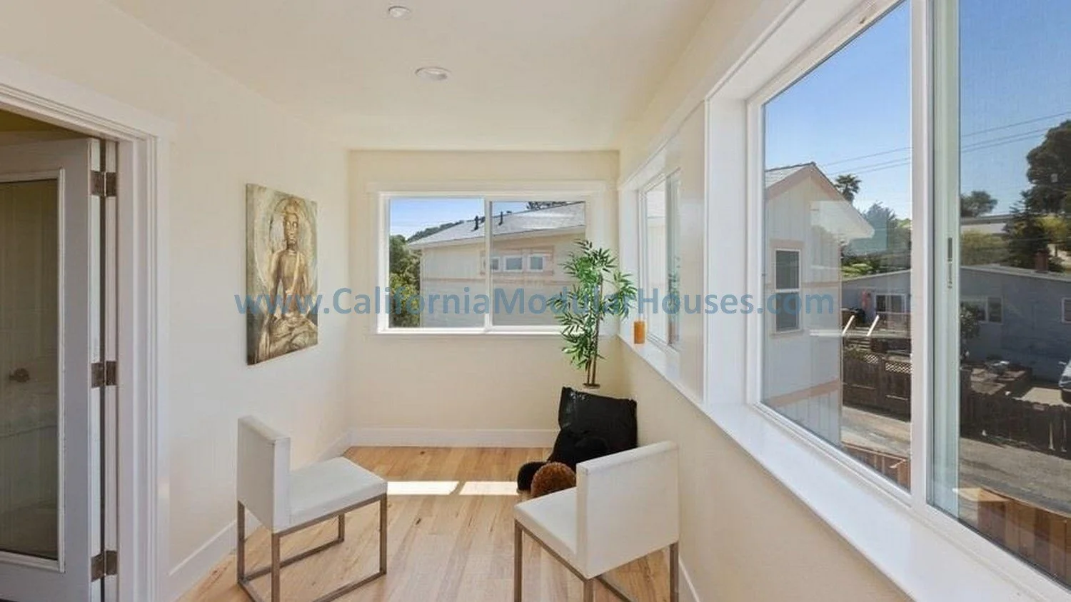 Sunlit enclosed porch with white walls, hardwood flooring, a potted plant, a framed art piece, and two white chairs near large windows showing neighboring houses and blue sky.  California modular home.  prefab modular CA