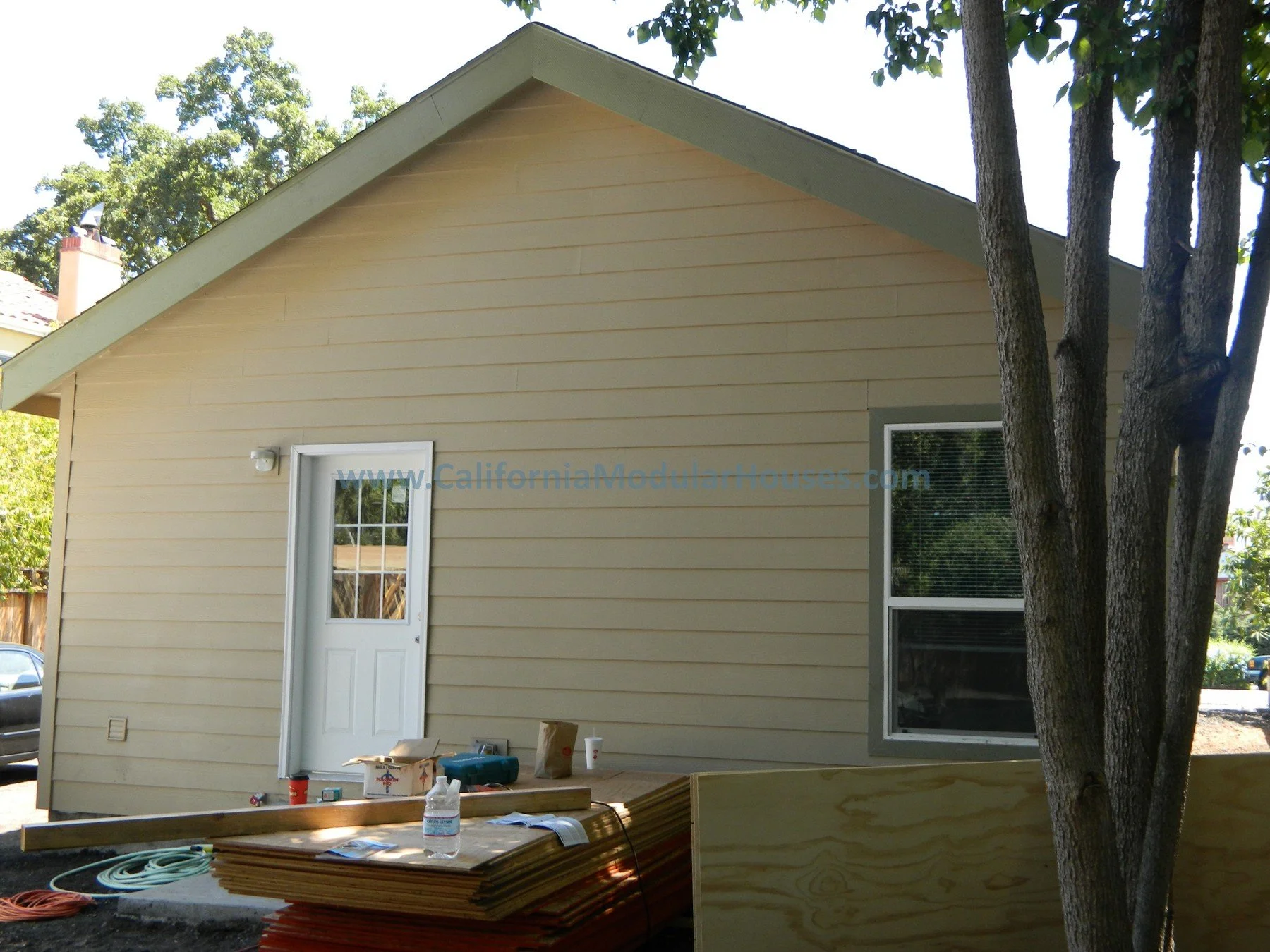 Side view of a beige house under construction with a door, a window, and building materials in the foreground, surrounded by trees.