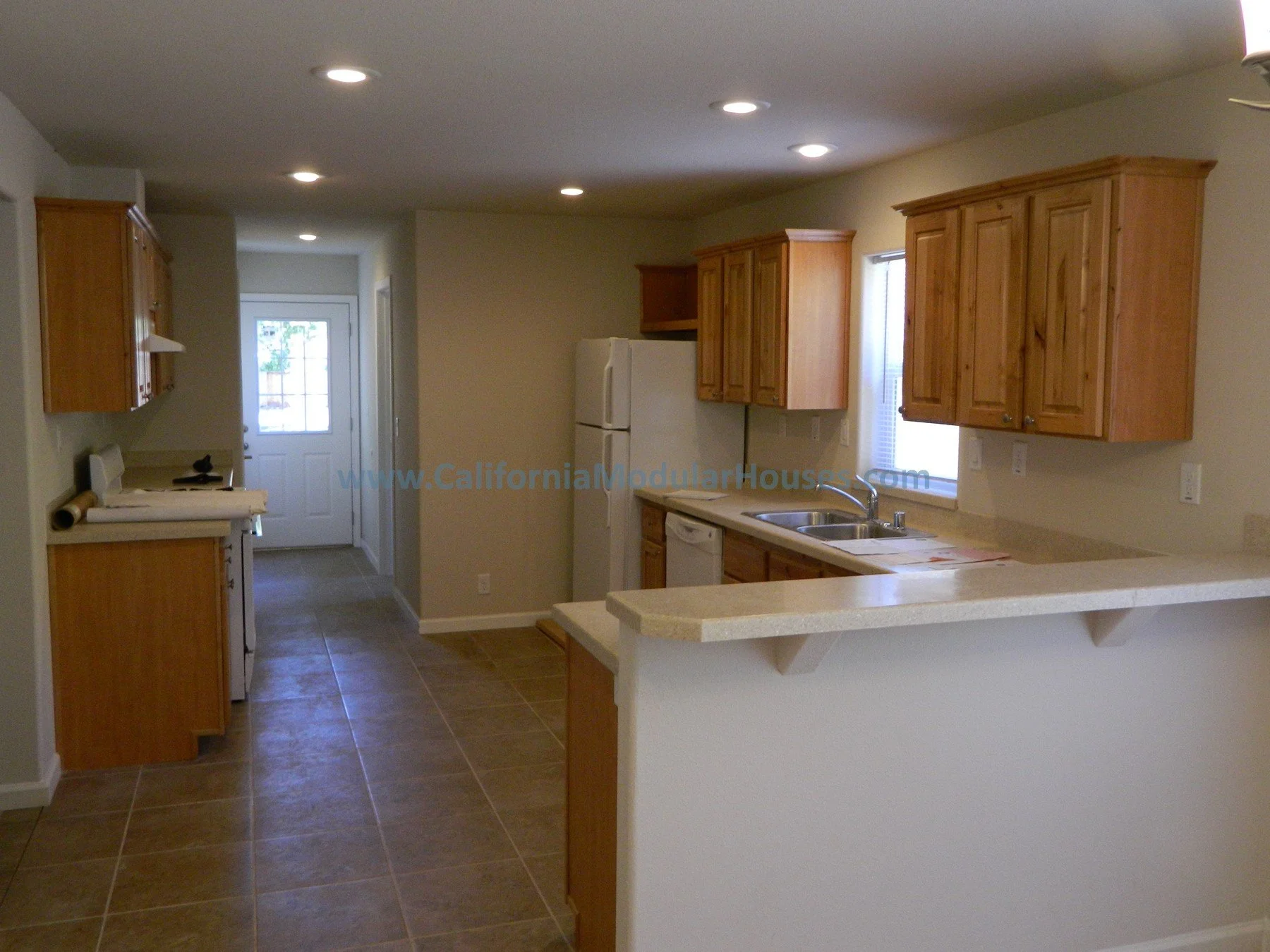 Kitchen with wooden cabinets, white appliances, beige countertops, a double sink, and tile flooring. A door with a window is visible in the background.