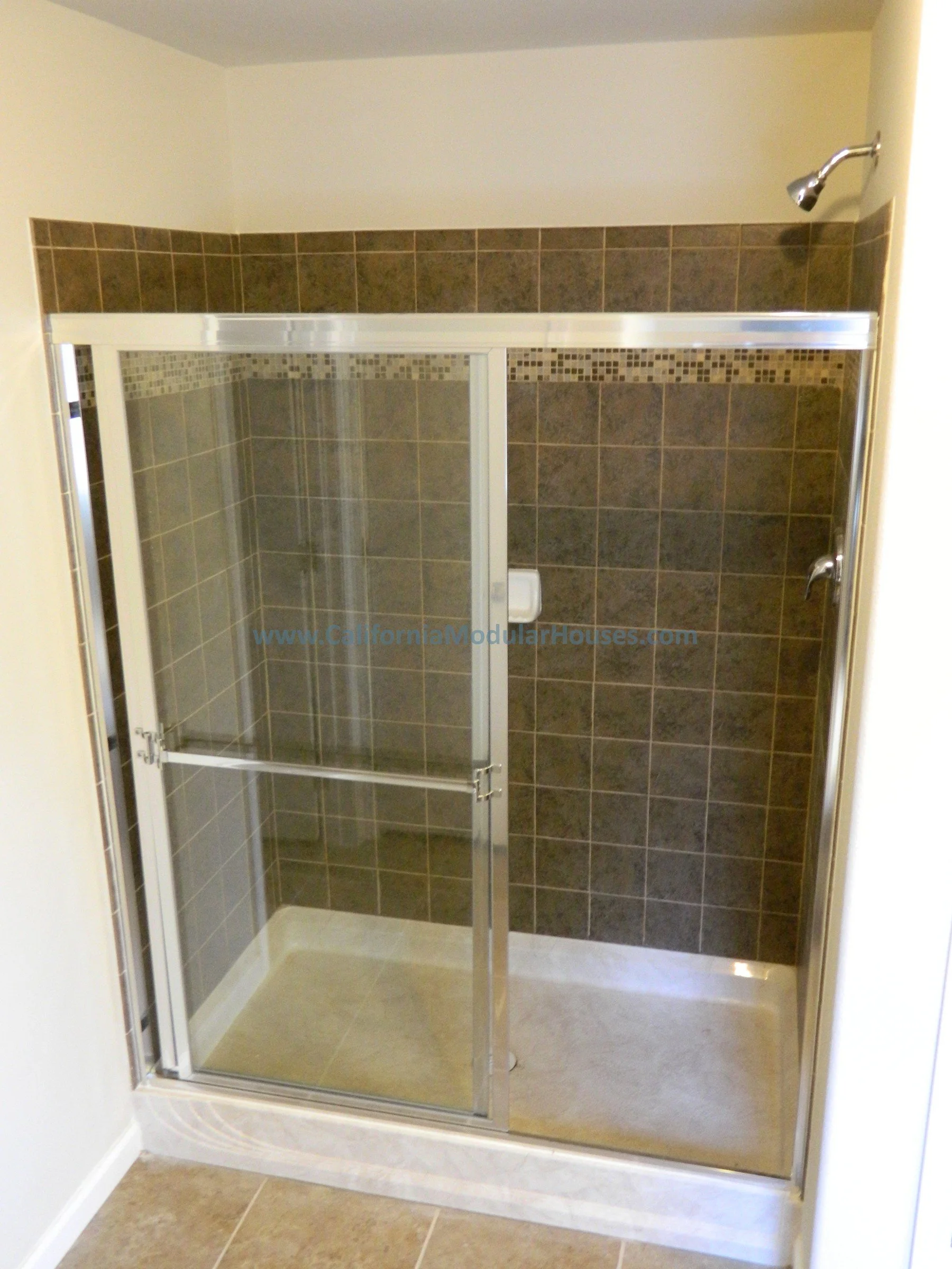 A walk-in shower with a clear glass sliding door, tiled brown walls, a small mosaic tile accent strip, and beige flooring.