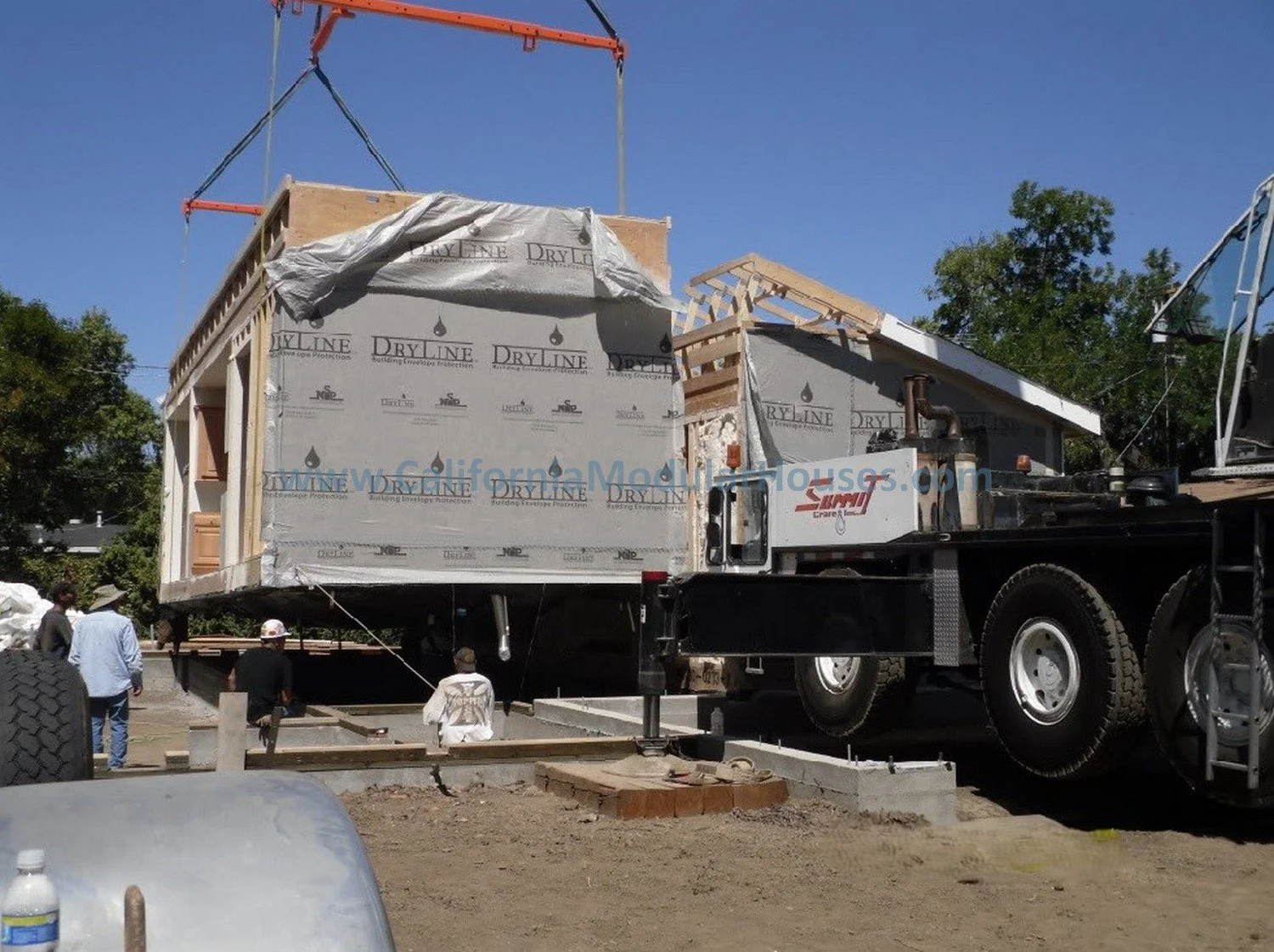 Construction site with a prefab modular house being built on a flatbed truck, workers on the ground, and a crane helping to position the structure.