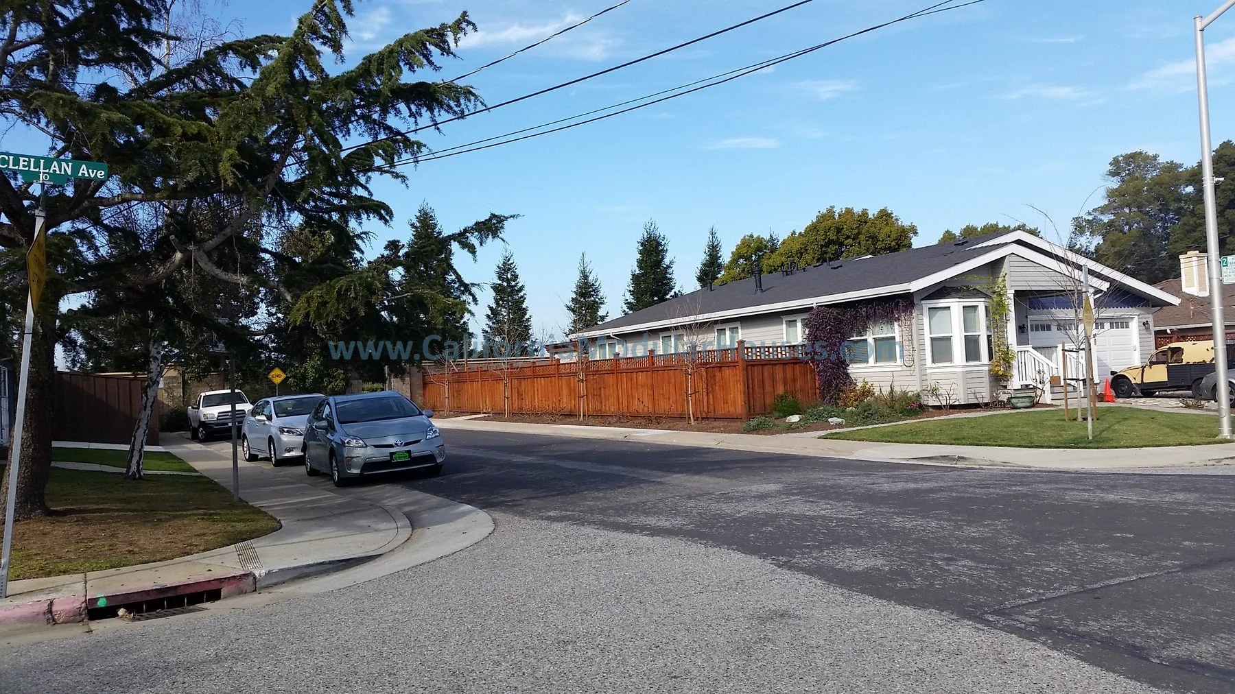 Residential street corner with stopped cars, a house with a fenced backyard, trees, and street signs. San Mateo County Modular, Prefab Home California,