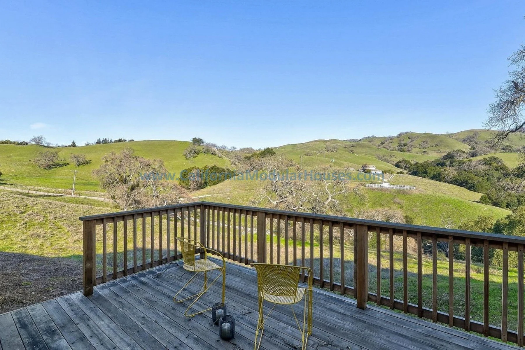 View of rolling green hills from a wooden deck with two yellow chairs and two small kettlebells.  Bay Area Prefab Modular Home, Prefab Modular Homes California.