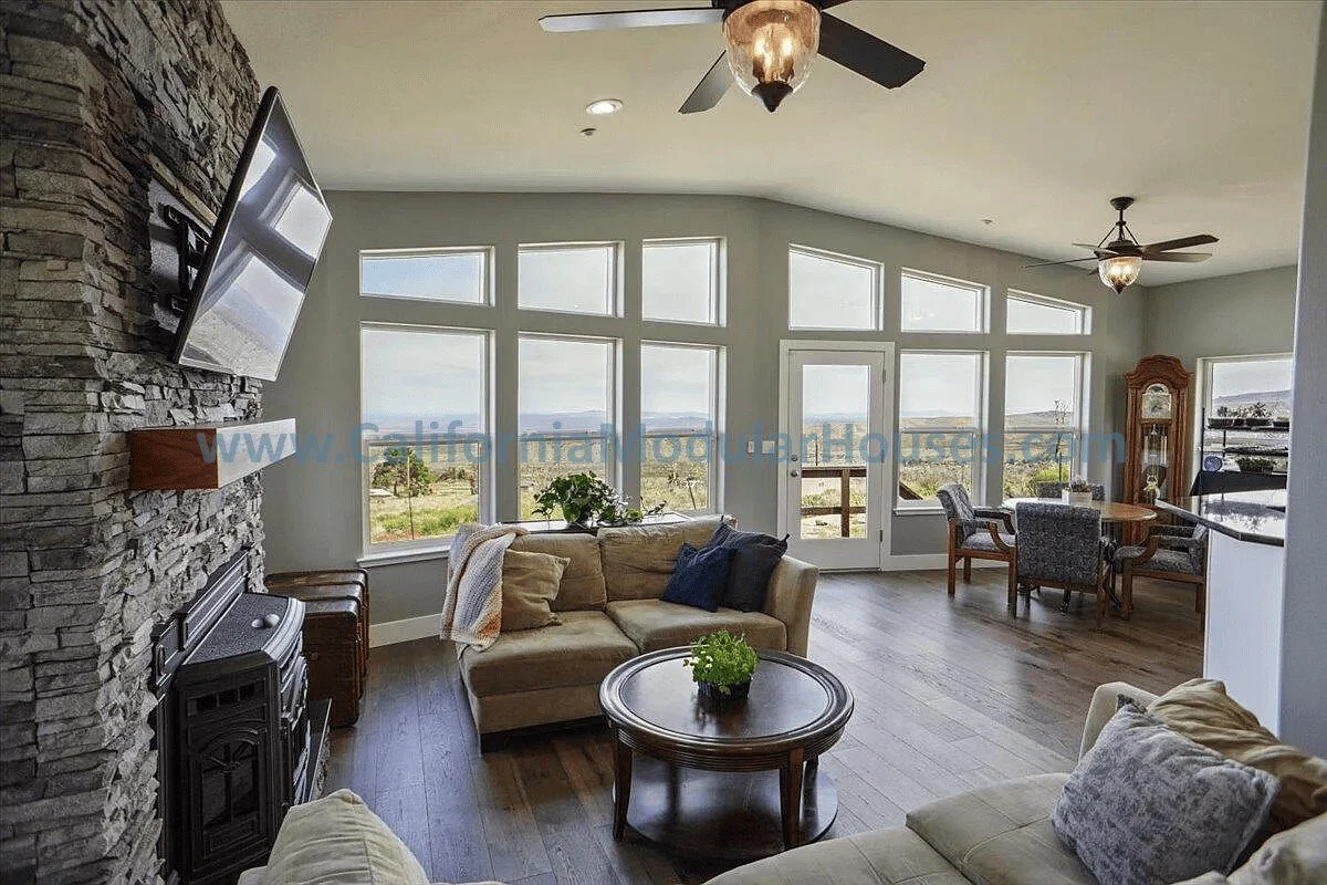 Living room with large windows, stone fireplace with mounted TV, beige sofas, wooden coffee table with a plant, dining area with round table and chairs, and a grandfather clock in the corner.