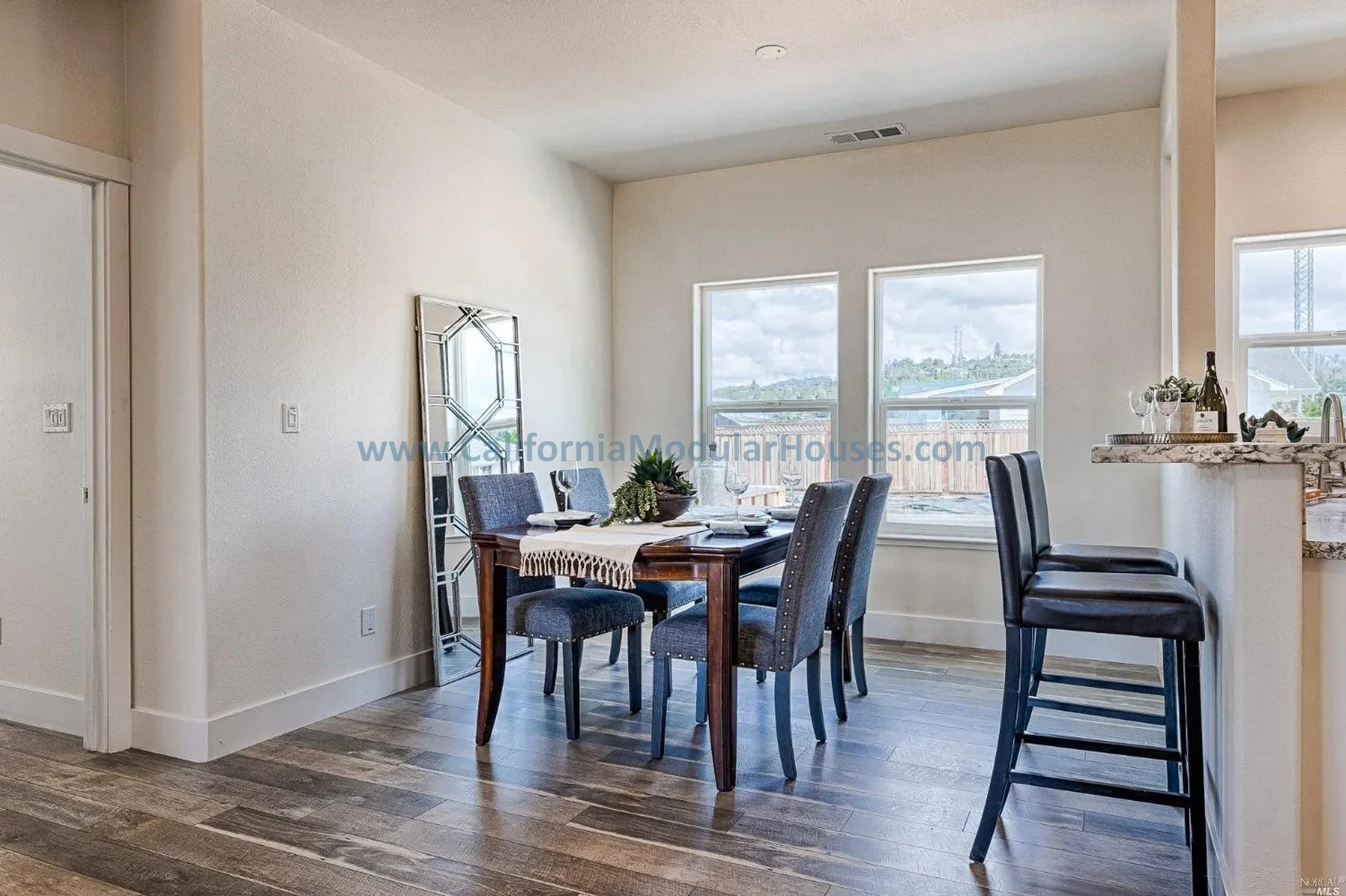 Dining area with a wooden table set with plates, wine glasses, and a centerpiece, surrounded by six upholstered chairs, situated near large windows overlooking a backyard with a wooden fence and cloudy sky.