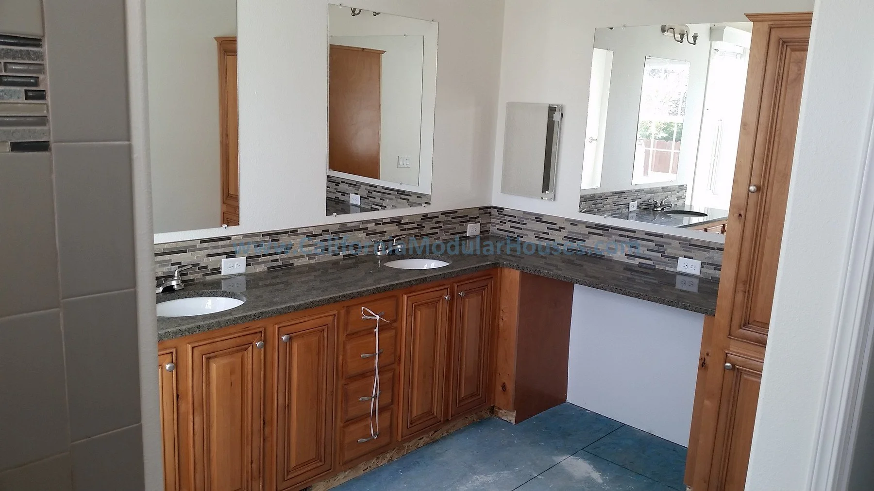Bathroom vanity with two sinks, wooden cabinets, gray granite countertop, and a large mirror. Part of the wall has a decorative tile backsplash. The background shows part of a room with windows and a door.  San Mateo County Modular.